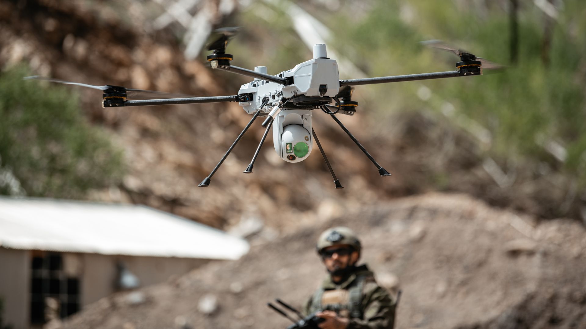White quadcopter drone with camera equipment flying outdoors in front of blurred background with soldier in camouflage holding a rifle.