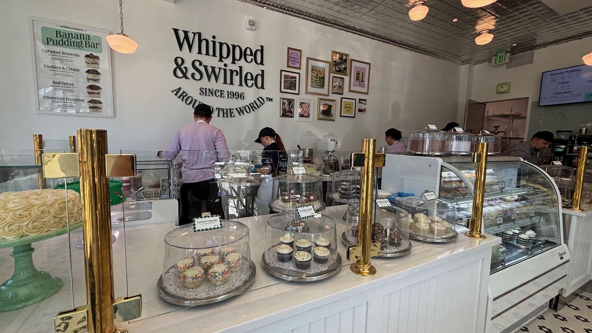 Inside Whipped & Swirled bakery: white walls, a large black sign, and framed photos. Glass display cases show cupcakes and cakes as staff and customers work behind the counter.