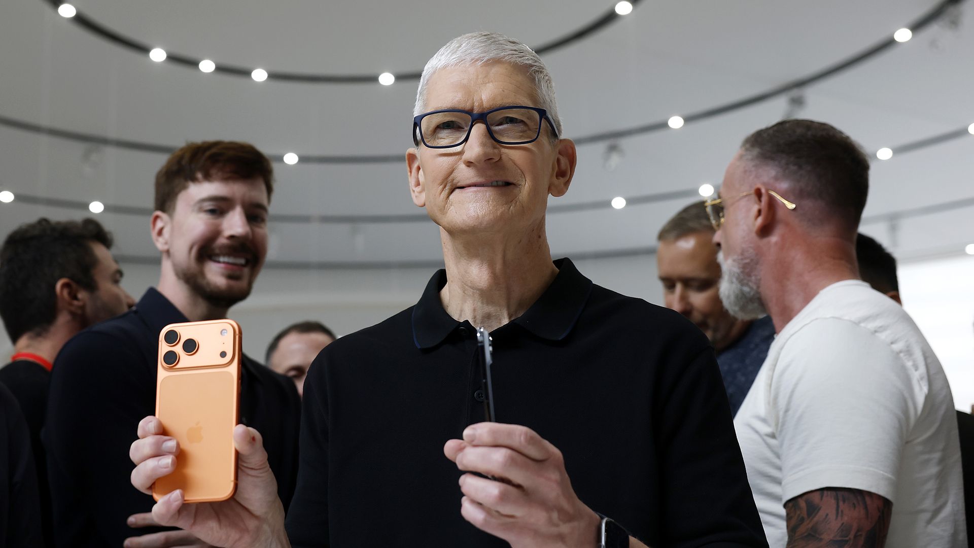 An man smiles while holding an orange iPhone and a pen-like device, surrounded by a small crowd in a bright modern space with circular ceiling lights.