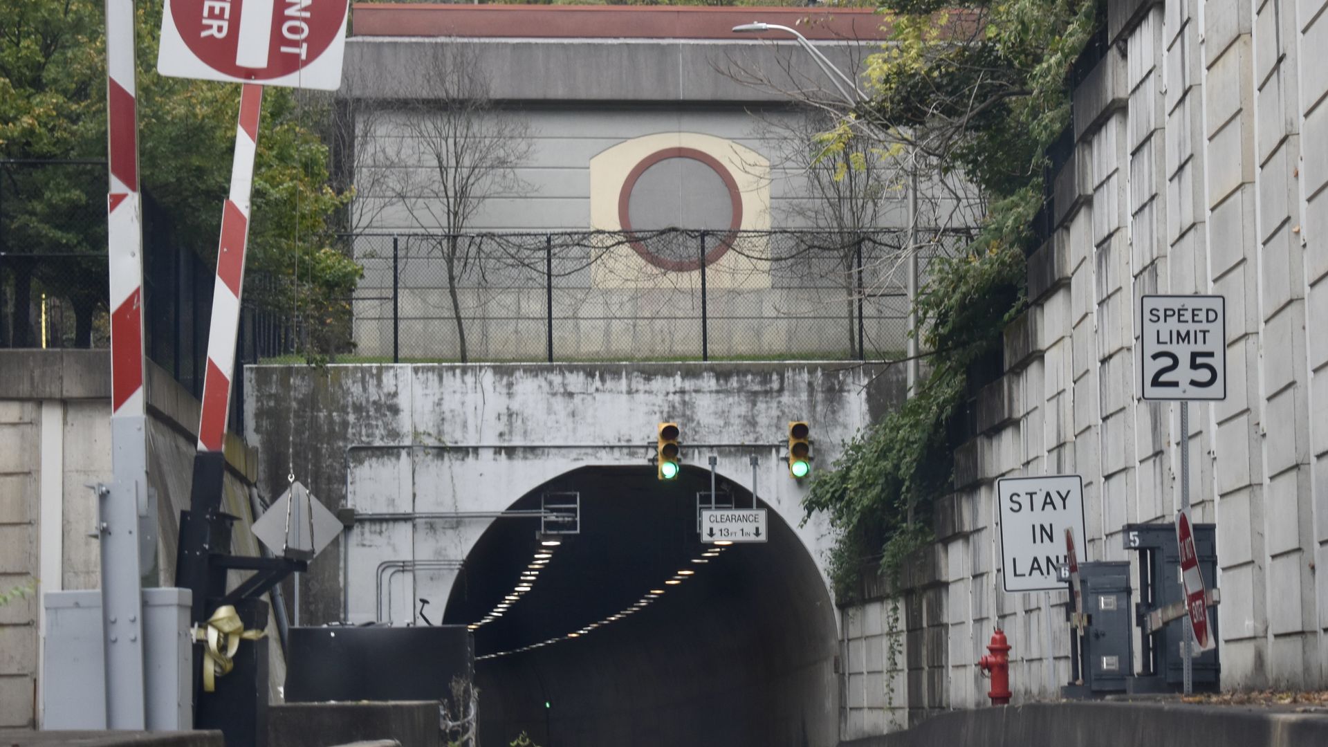 Urban tunnel entrance seen head-on with red-and-white striped barriers on the left, a beige wall bearing a circular red target sign behind a chain-link fence, and green traffic lights with a 25 mph sign on the right.