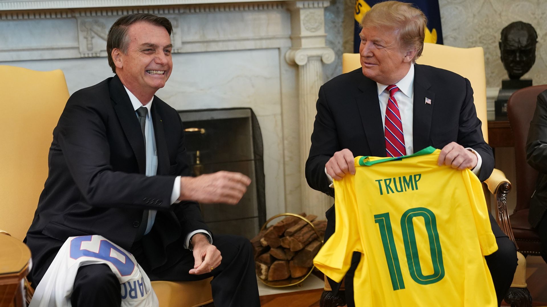 President Trump holds up a yellow sports jersey with the letter ten on it and his name, while sitting next to the Brazilian president in the Oval Office.