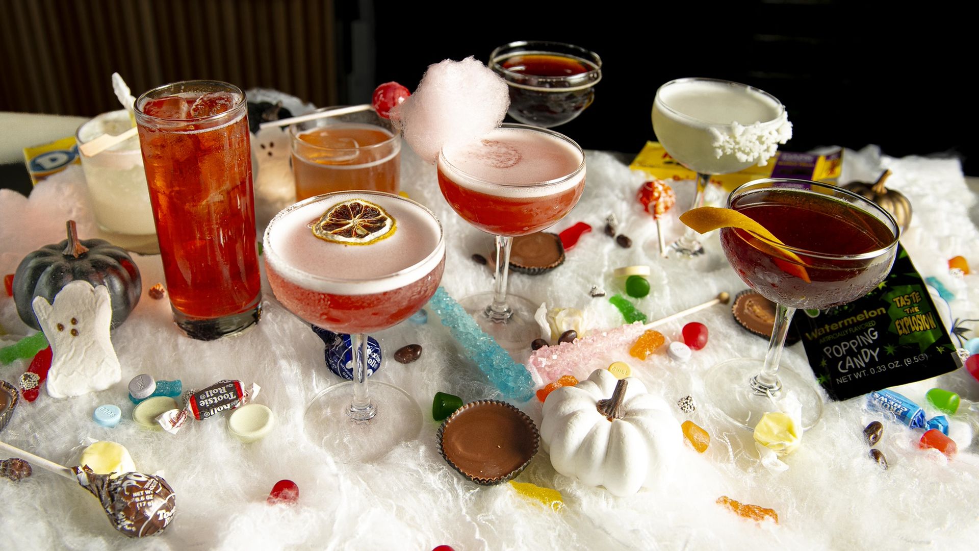 A festive Halloween-themed drink assortment on a white fluffy surface with small pumpkins, candy, and cocktails garnished with dried citrus, cotton candy, and citrus peel.