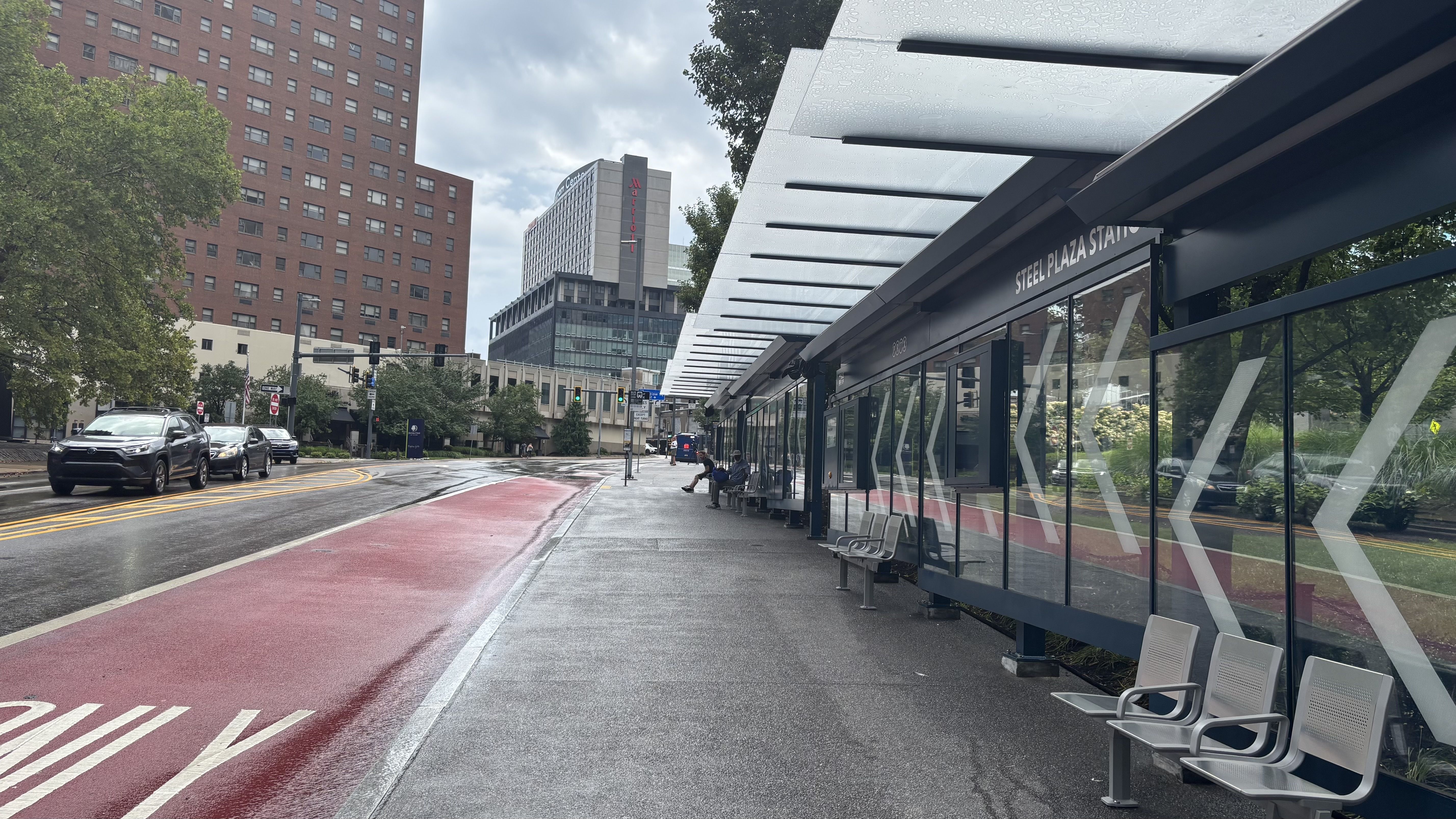 View of Steel Plaza Station bus stop with glass walls and metal benches on a wet gray day, red bus lane, traffic lights, cars, trees, and tall buildings in the background.
