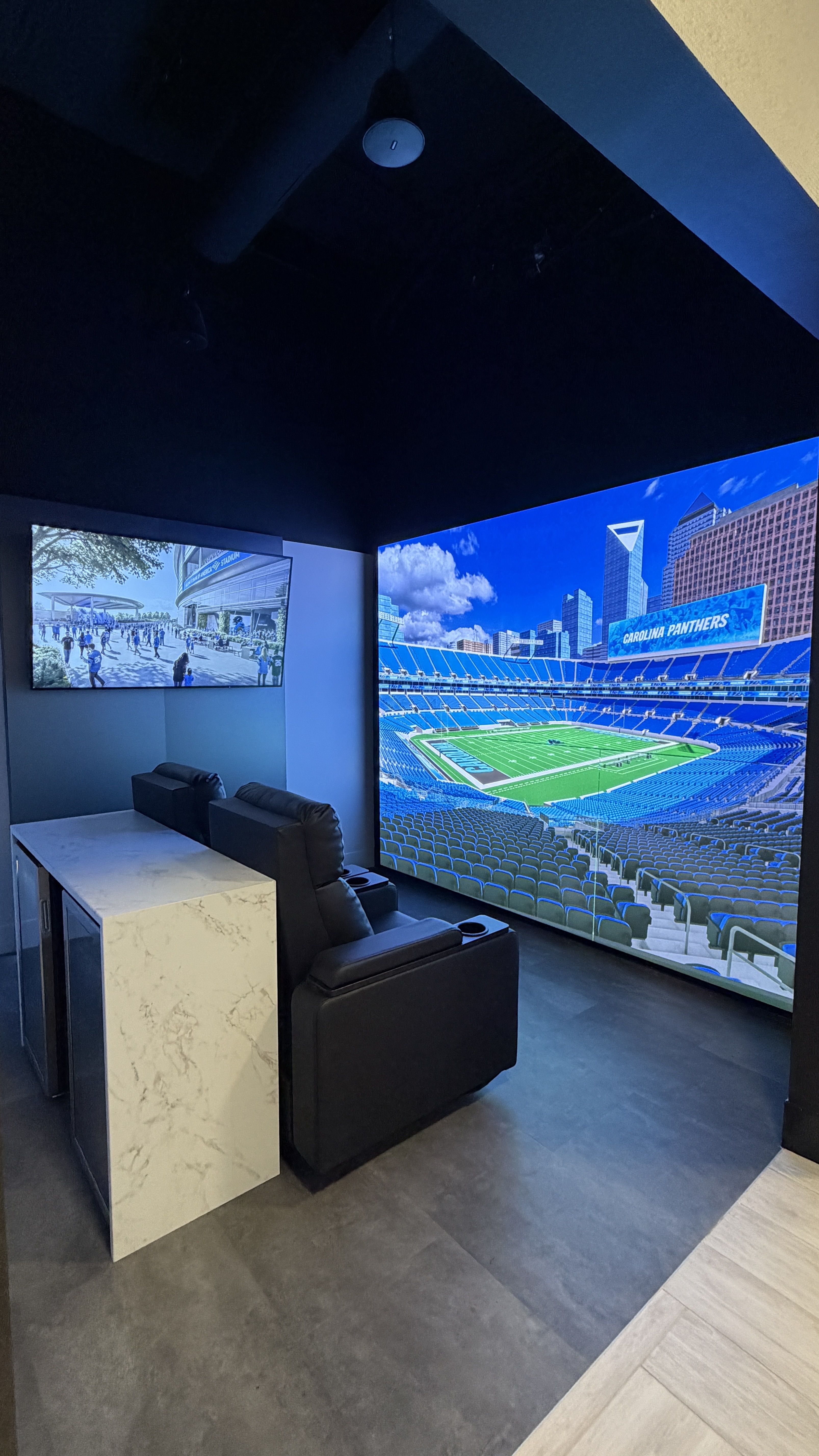 Dark private viewing room with two black leather recliners facing a large wall screen of a Carolina Panthers stadium; a smaller TV shows the exterior stadium, with blue lighting and a white marble counter.