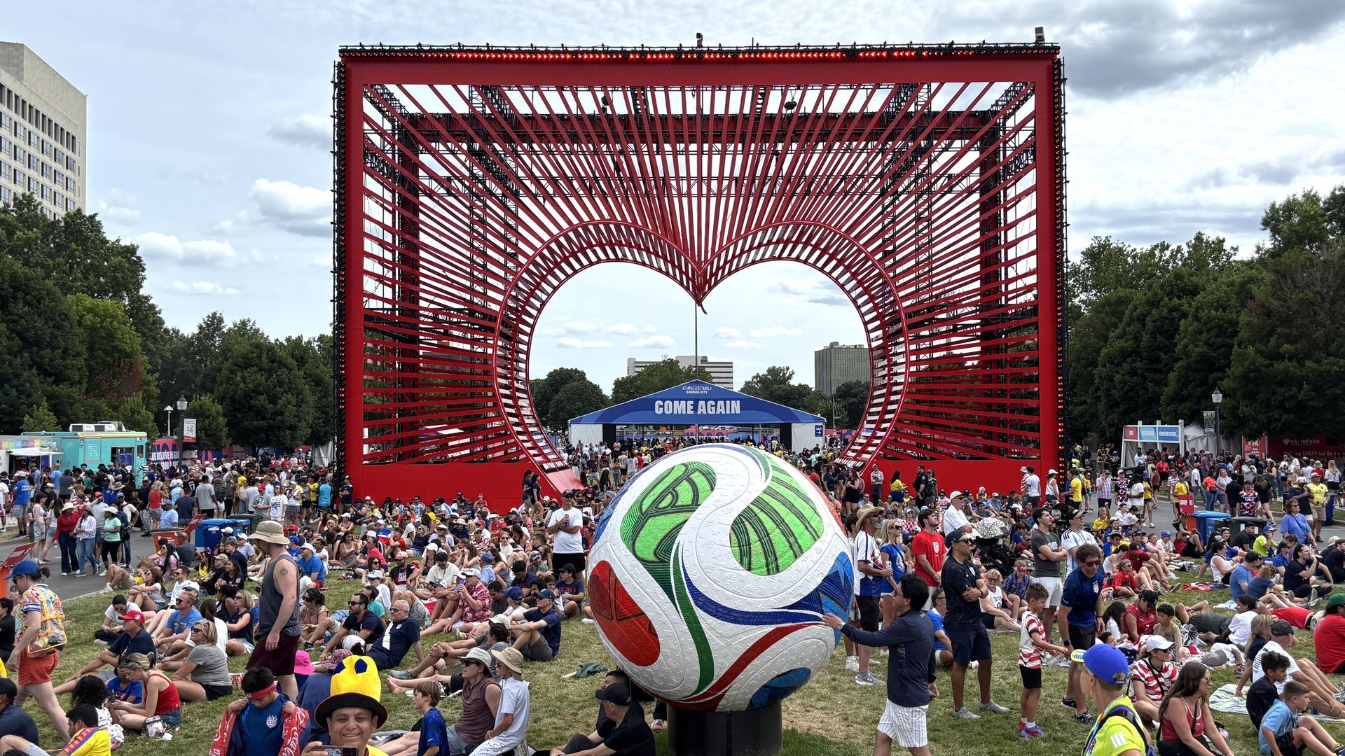 Crowd at an outdoor festival gathered around a large red geometric sculpture forming a heart; a blue tent reads "COME AGAIN" behind as trees and city buildings rise beyond.
