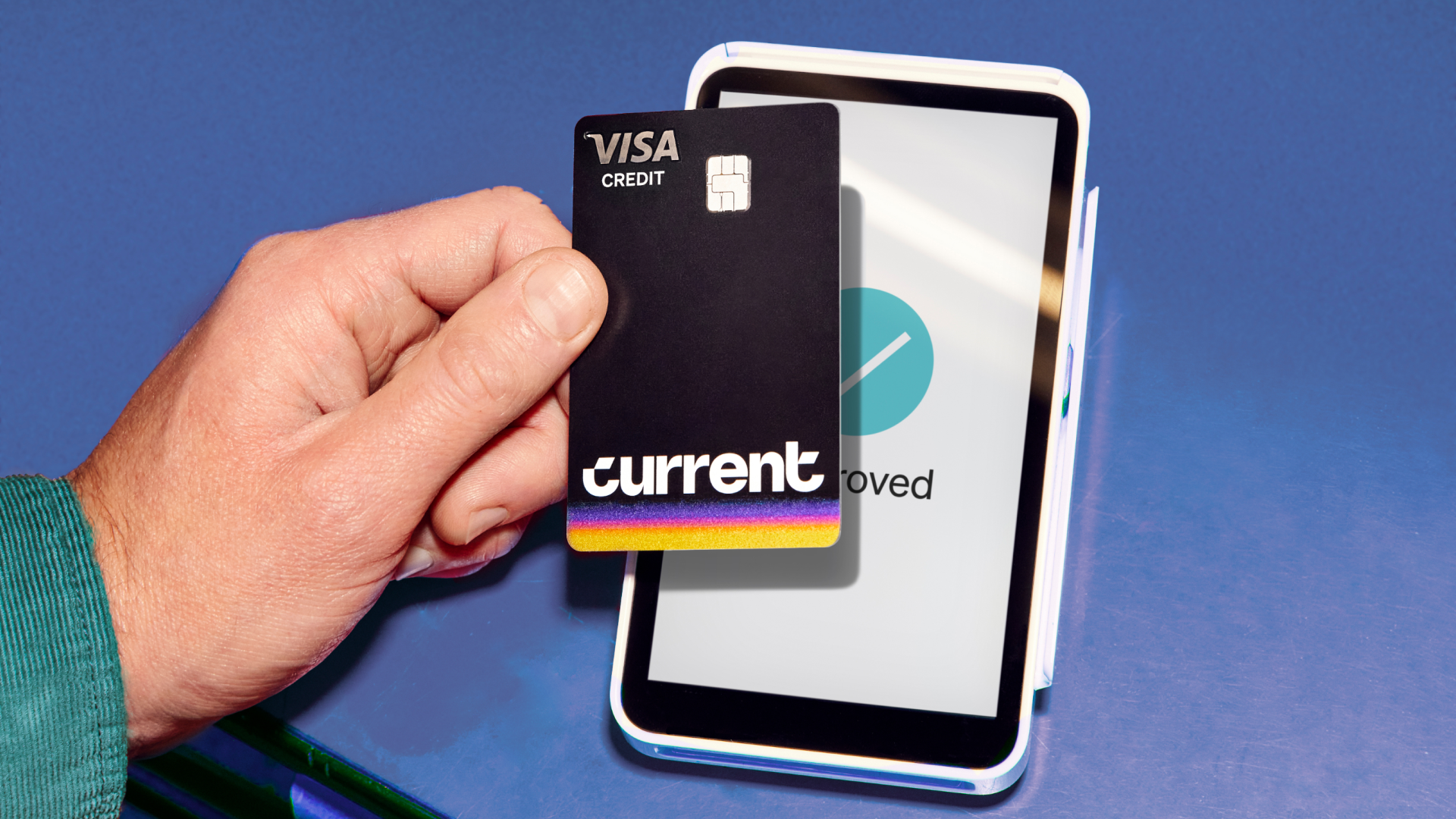 Hand holding a black Visa credit card labeled "Current" with a rainbow stripe, near a white card reader showing a checkmark and the word "approved" on a blue surface.