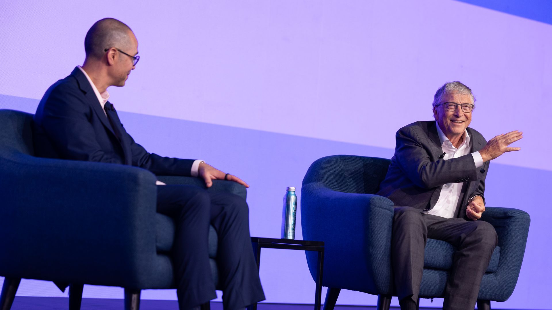 Breakthrough Energy executive director Rodi Guidero talks with co-founder Bill Gates on stage at the Breakthrough Energy Summit in London. Both sit on blue armchairs, with a screen behind them displaying purple and blue blocks of color.