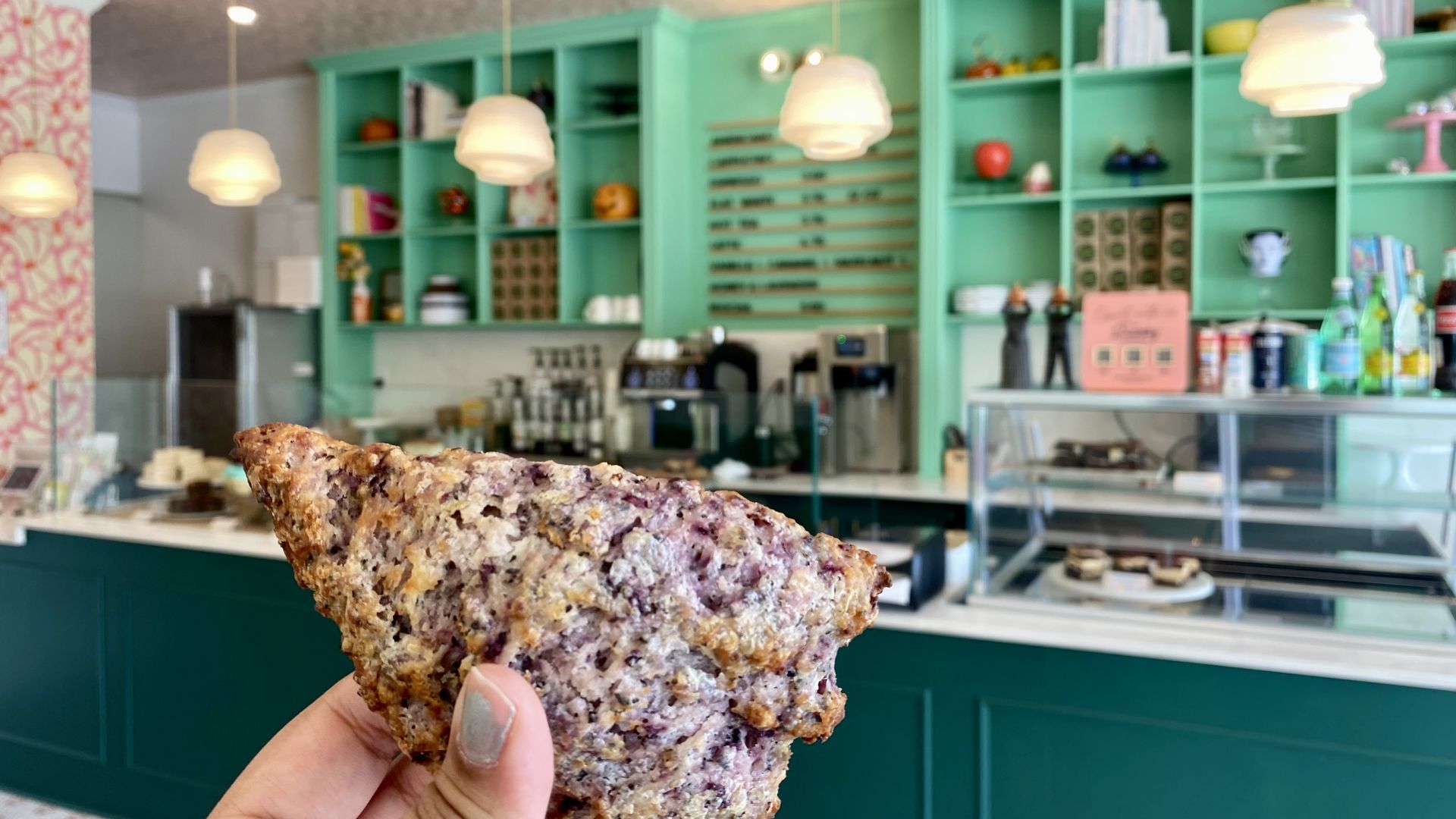 Hand holding a triangular earl grey blackberry scone inside a cafe with teal shelves, hanging white pendant lights, and a glass display case with pastries.