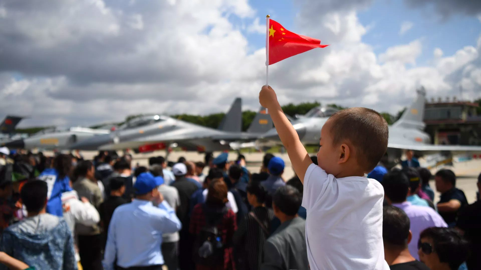 A little Chinese boy hoding a Chinese flag at an airbase surrounded by people.