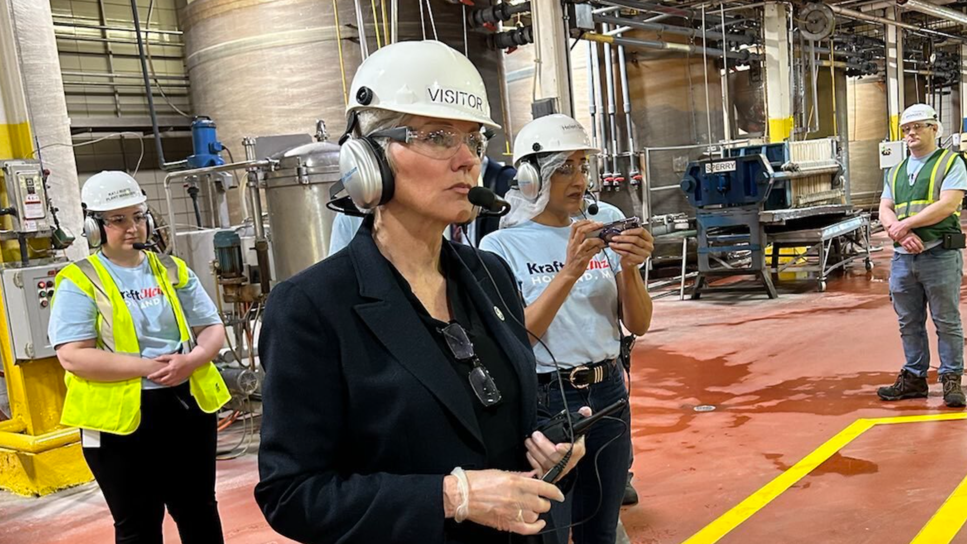 Jennifer Granholm is standing on a factory floor, wearing a hard hat and a headset.