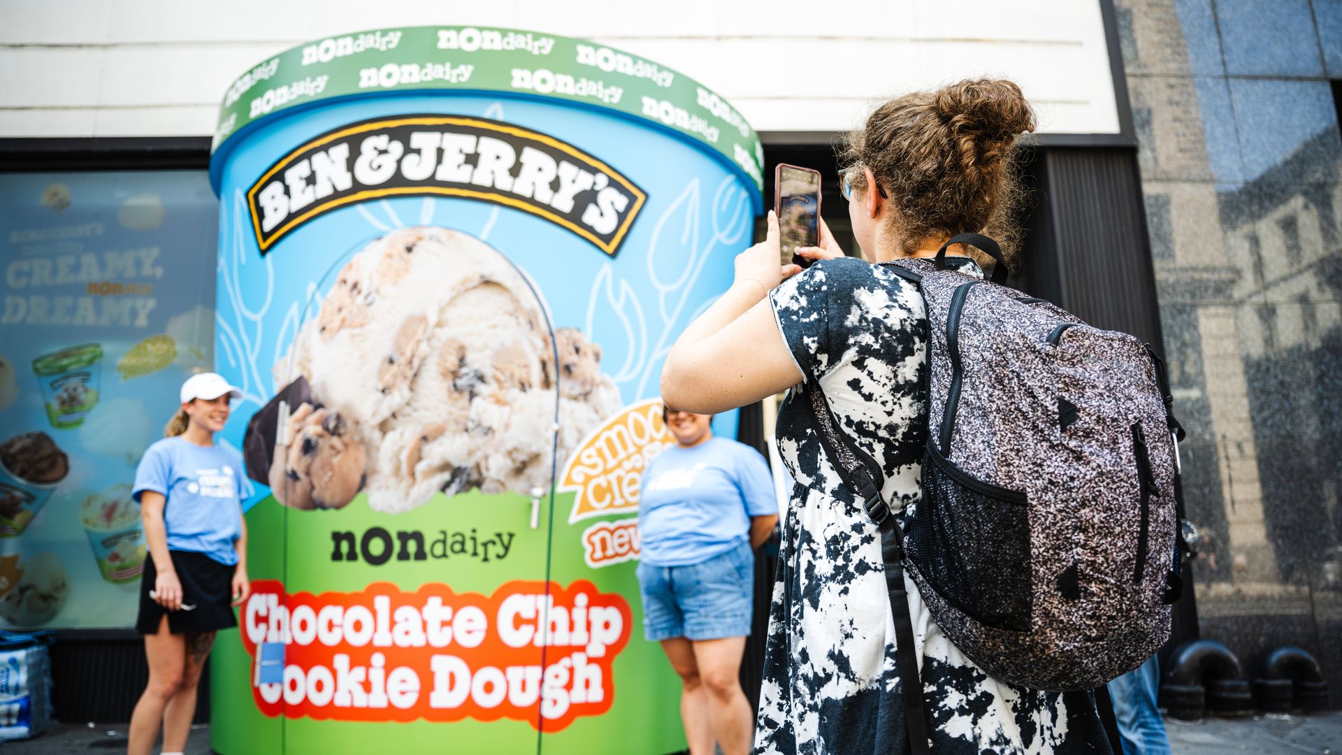 Someone takes a photo of two people posing in front of a giant pint of Ben and Jerry's non dairy chocolate chip cookie dough