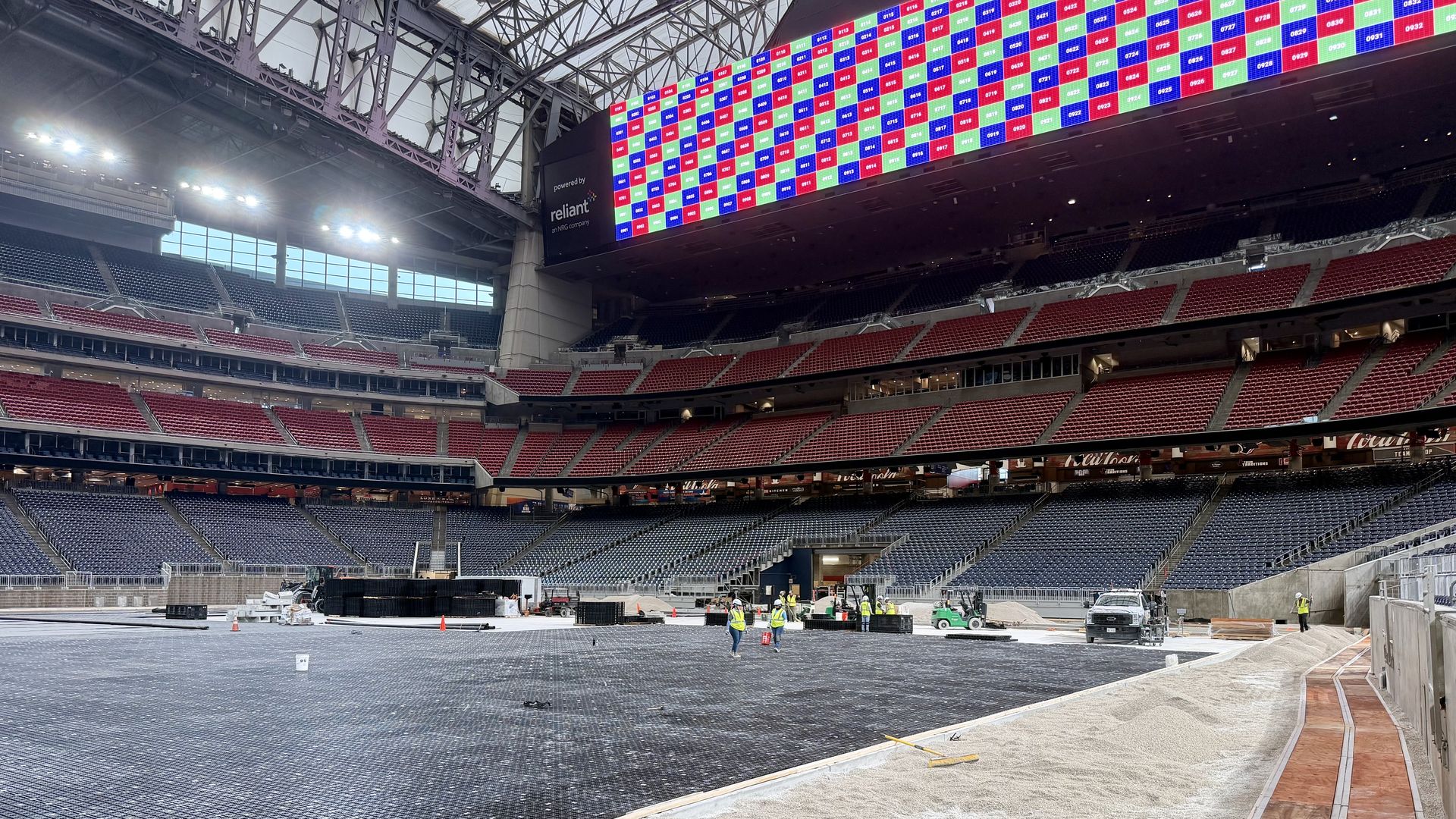 Interior of a large stadium under construction: steel framework overhead, bright multi-colored video board spanning stands, red seating, and workers in yellow vests on a partially finished floor.