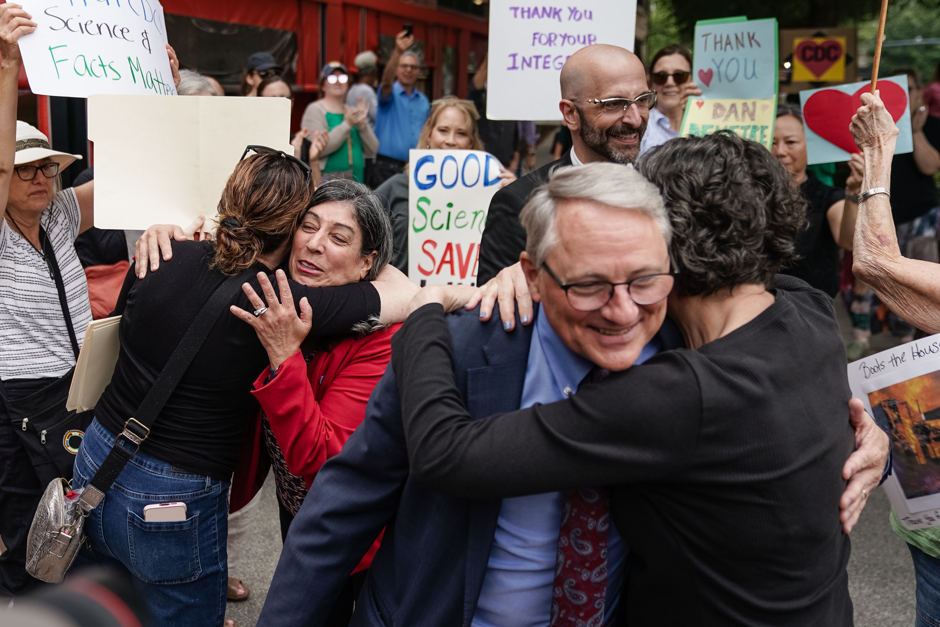 A group of people hugging and smiling at an outdoor rally with colorful signs praising science, facts, and integrity in the background. Some hold signs with hearts and CDC logos.