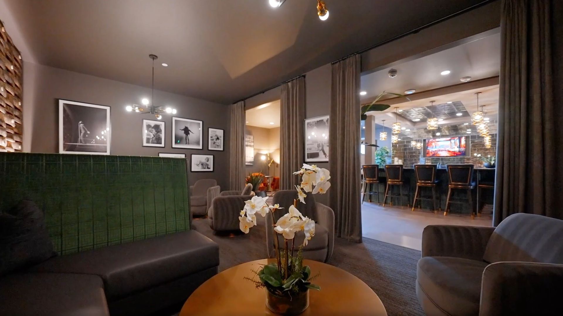 Cozy lounge with gray walls and curtains, a wall of black-and-white photos, gray seating, and a round table with white orchids. A green-tiled partition and a bar with stools in the background.