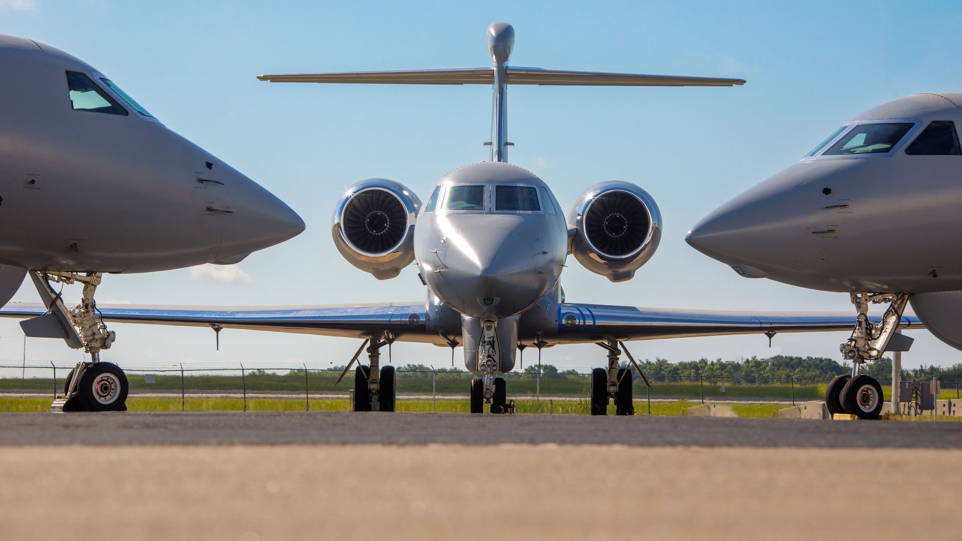 Front view of a silver jet with two jet engines, parked on a runway between the noses of two other similar jets, under a clear blue sky.