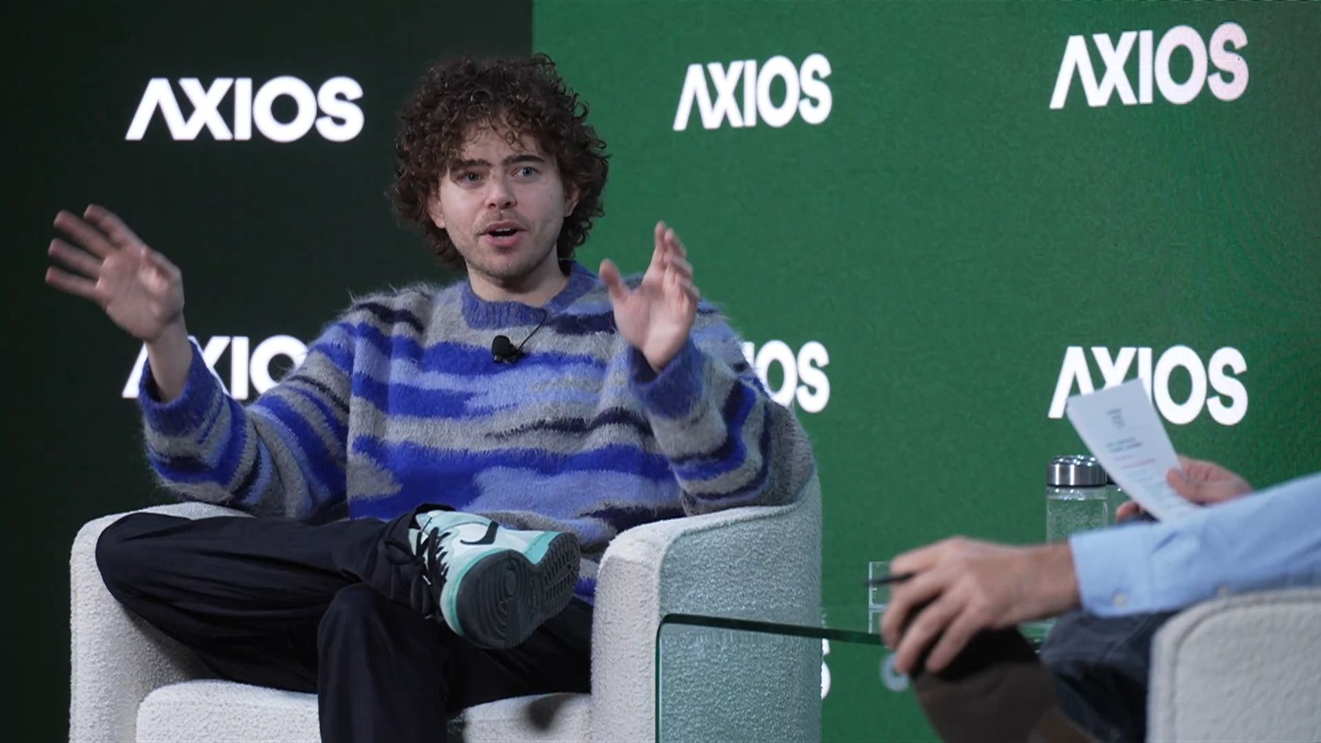 A man with curly hair wearing a blue and gray striped fuzzy sweater and mint green sneakers speaks while sitting in a white armchair on a stage with green background displaying "AXIOS" logos.