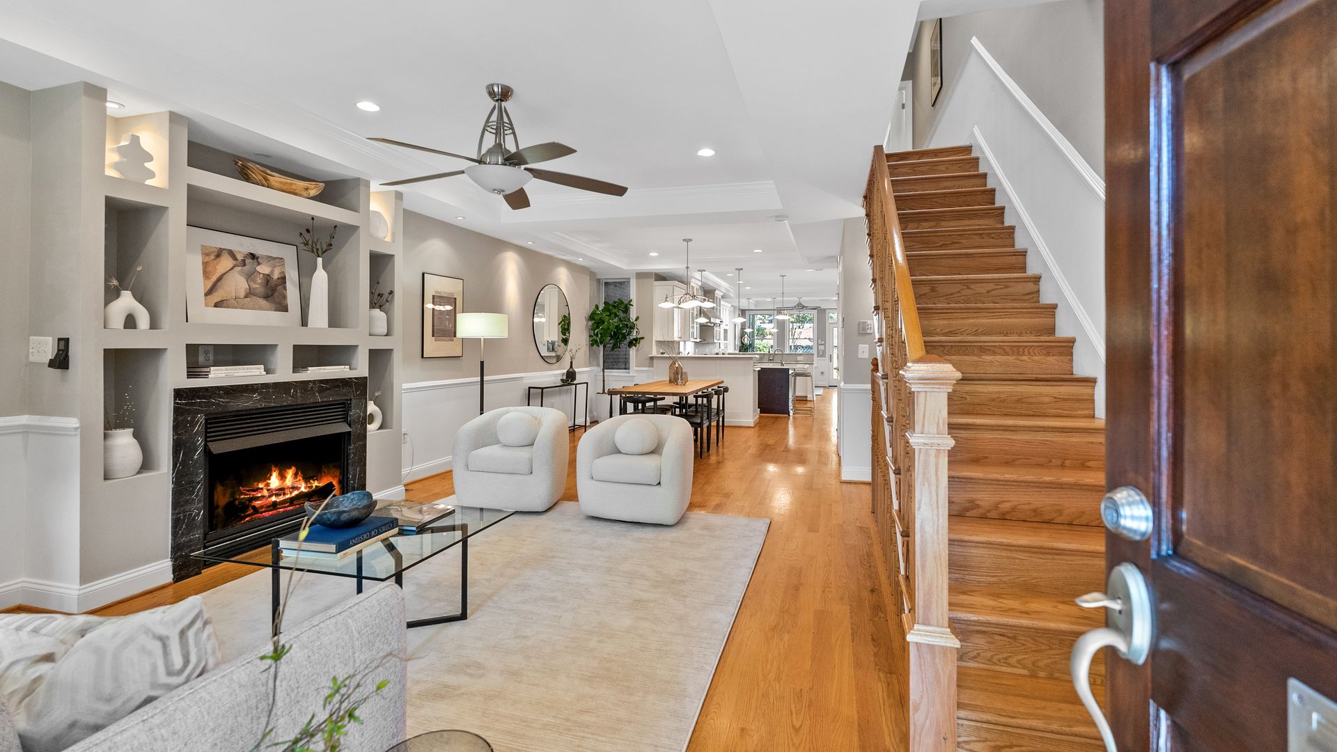Open living room with beige walls, wooden floor, lit fireplace, two white armchairs on gray rug, glass coffee table, wooden dining table, and stairs to the right.
