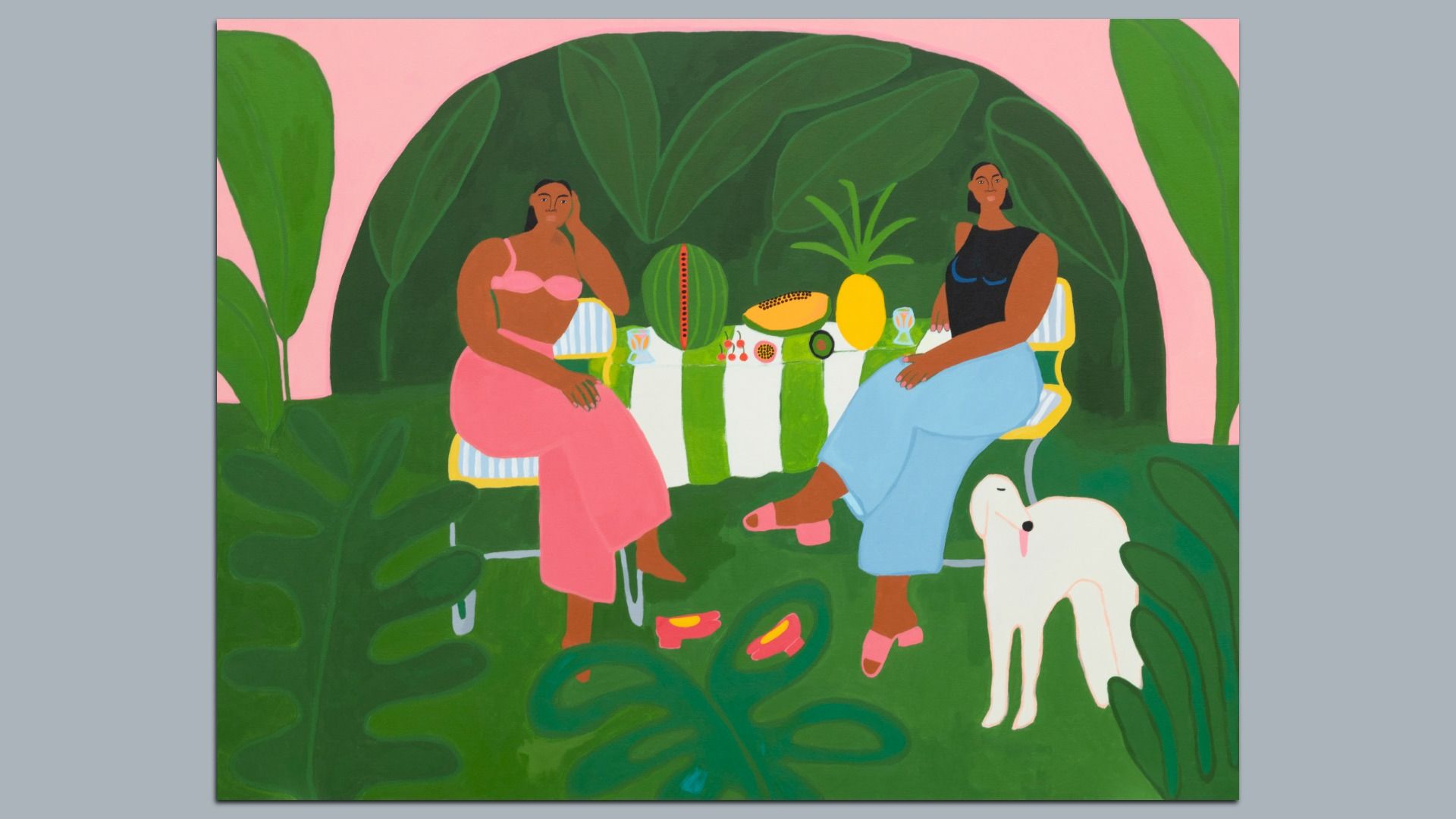 Two women sit at a striped table in a lush tropical garden; pink wall behind. They wear pink and blue outfits. Fruit (watermelon, pineapple) on the table; a white dog stands nearby.
