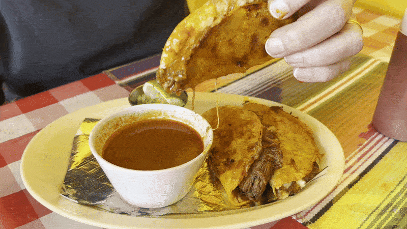 Image shows a hand dipping a birria taco in a bowl of consome.