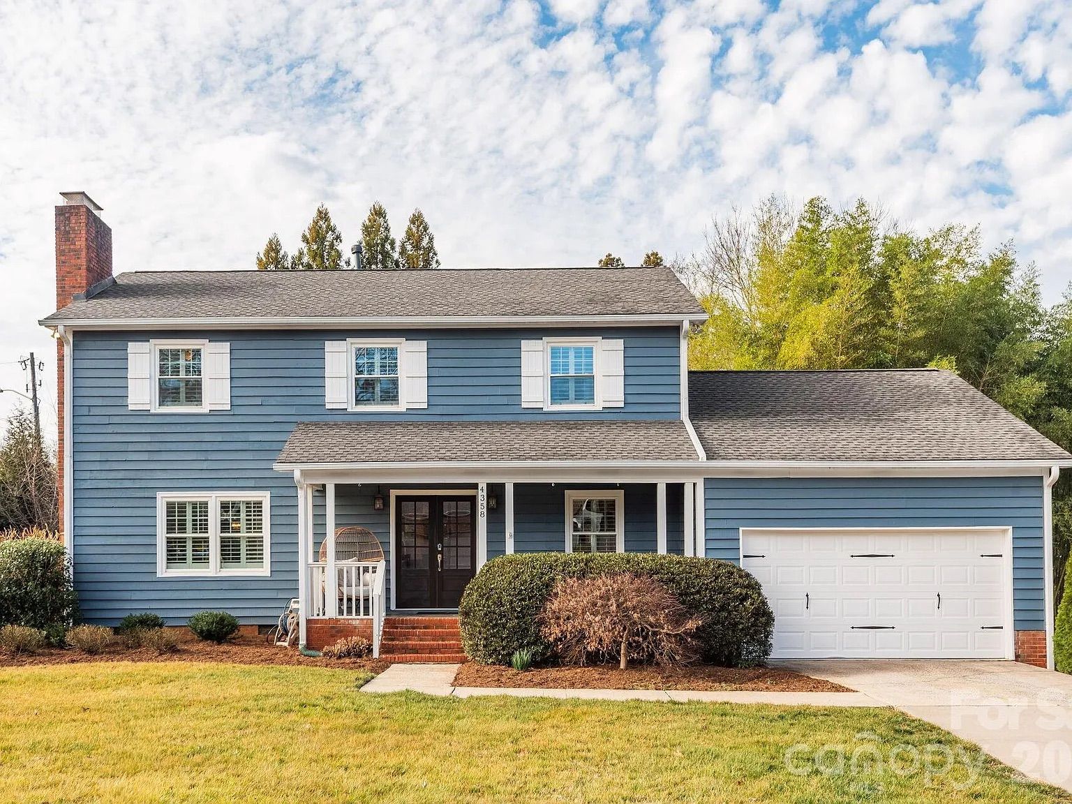 Two-story blue house with white shutters, brick chimney and steps, black front door, porch with hanging chair, attached white garage, green lawn, and partly cloudy sky.