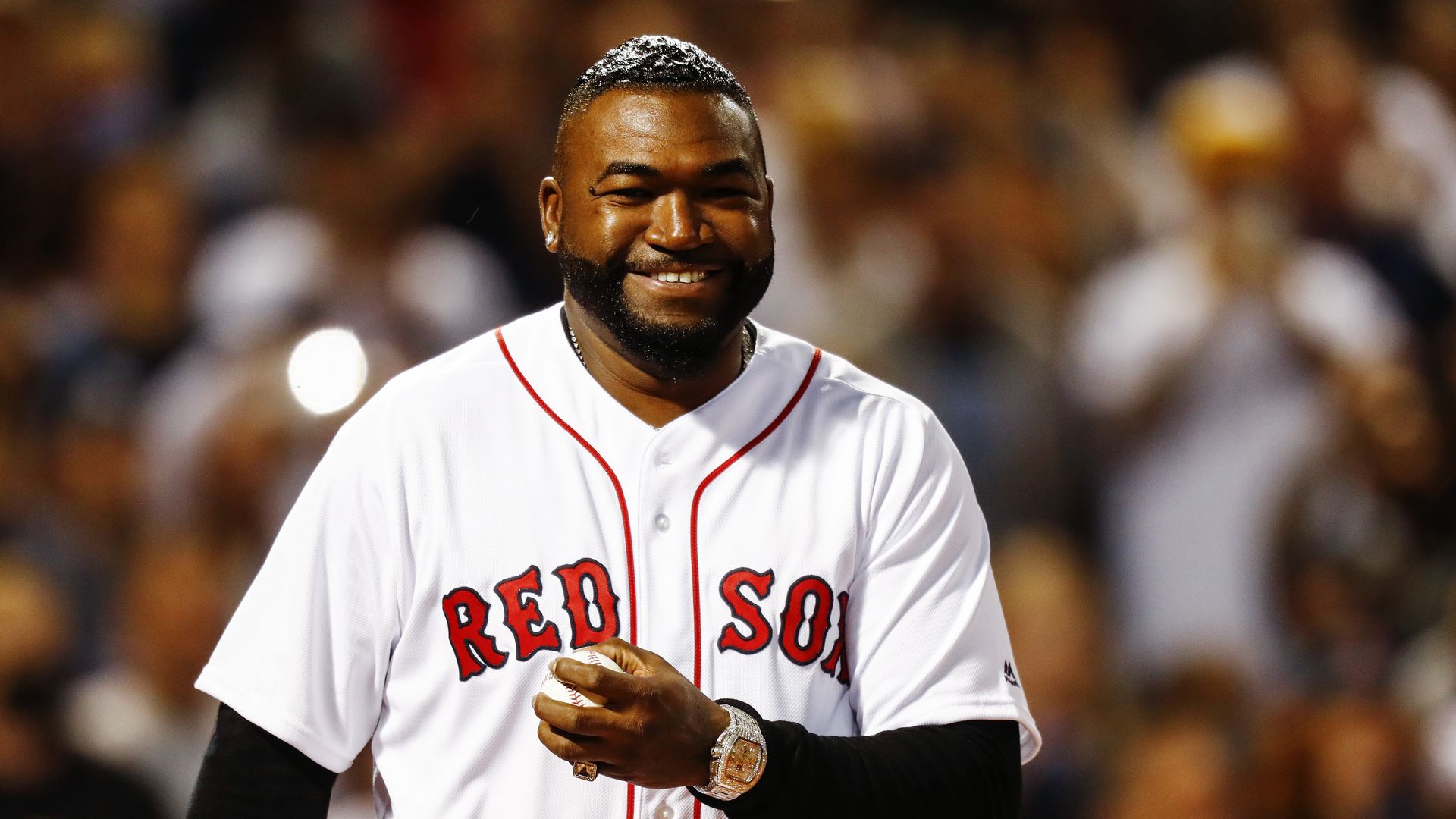Photo of David Ortiz in a white Red Sox uniform. He's standing in a stadium, grinning and holding a baseball in his left hand.