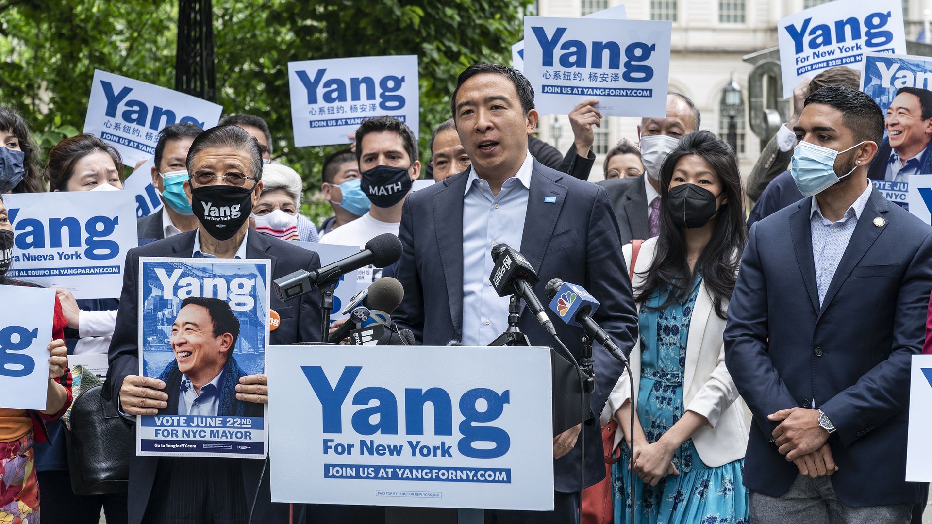 Photo of Andrew Yang speaking from a podium with a crowd of people holding signs supporting Yang behind him