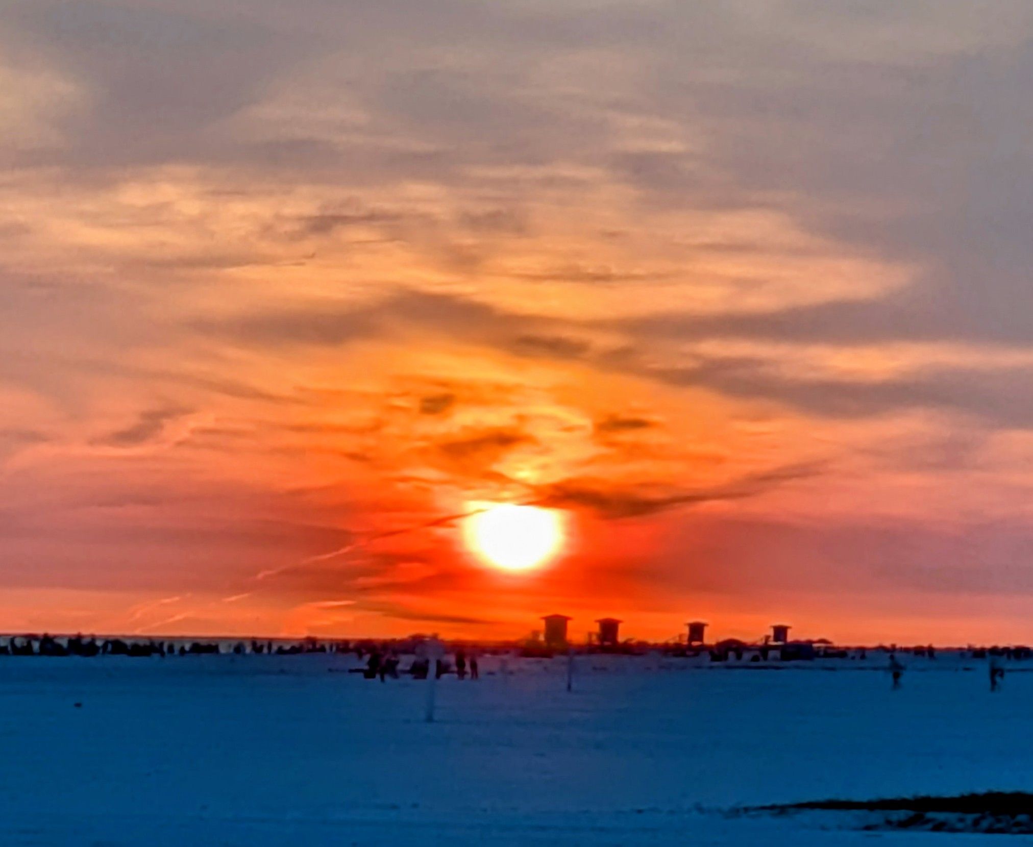 Blurry sunset over the ocean with an orange sun on the horizon, pink and blue sky, and silhouettes of people on the beach behind a row of posts along the shore.
