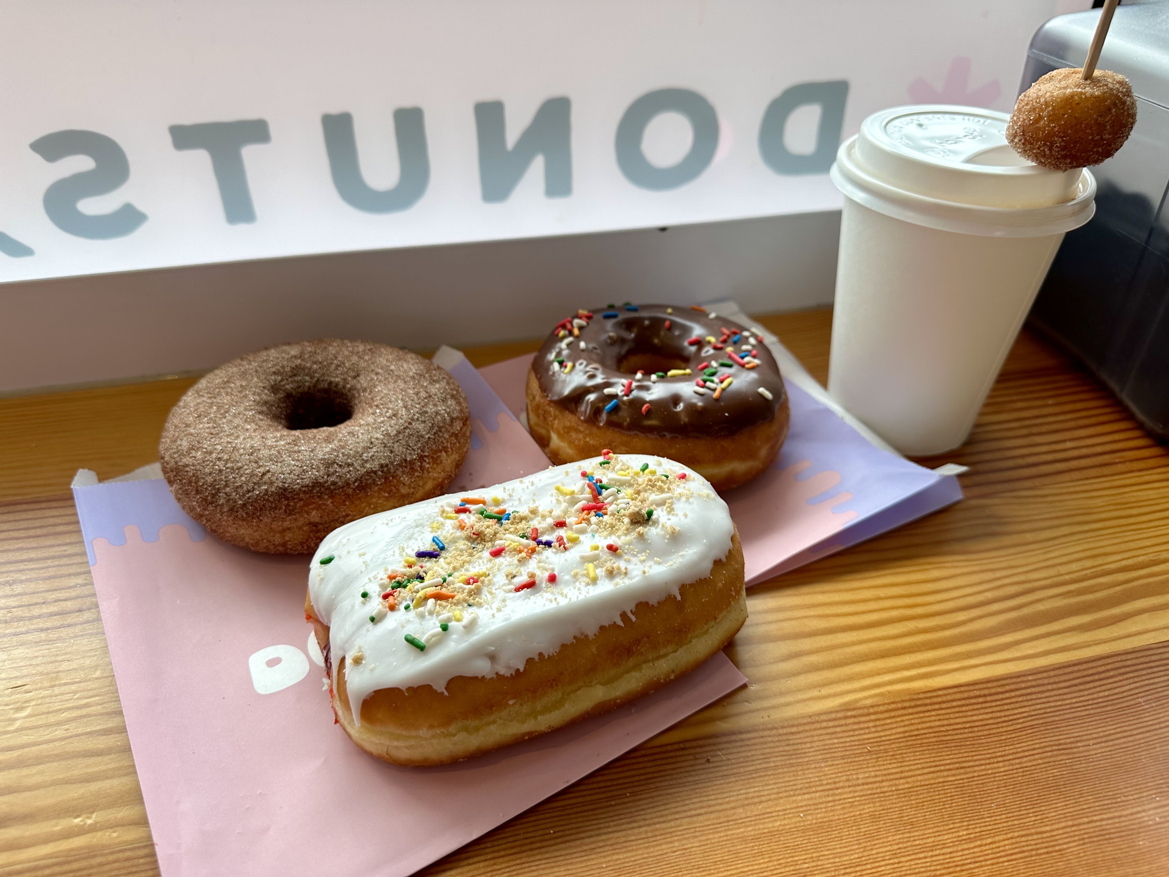 A cinnamon sugar and chocolate frosted doughnut behind a bar that looks like a pop-tart, next to a coffee with a  doughnut hole on the straw, on a wooden counter next to a window that says "donuts"