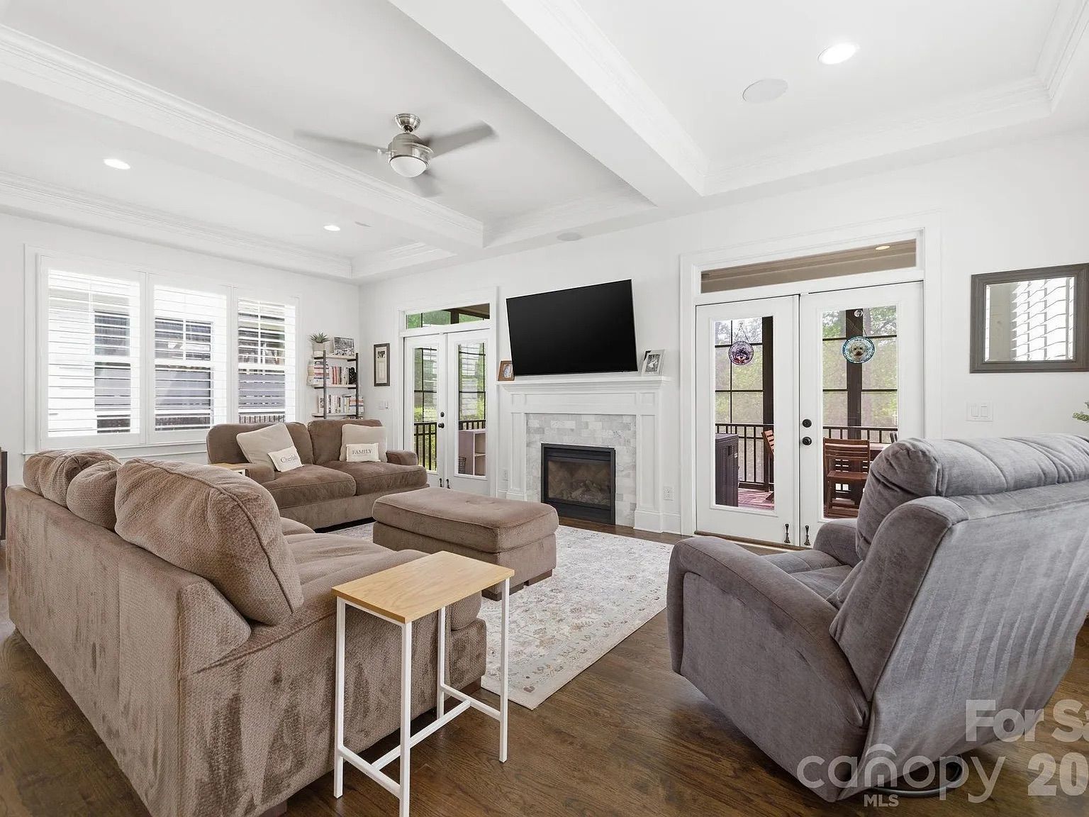 Bright living room with white walls, exposed beams, and a ceiling fan. A brown sectional and gray recliner face a stone fireplace with a mounted TV; French doors and blinds-filled windows let in daylight.
