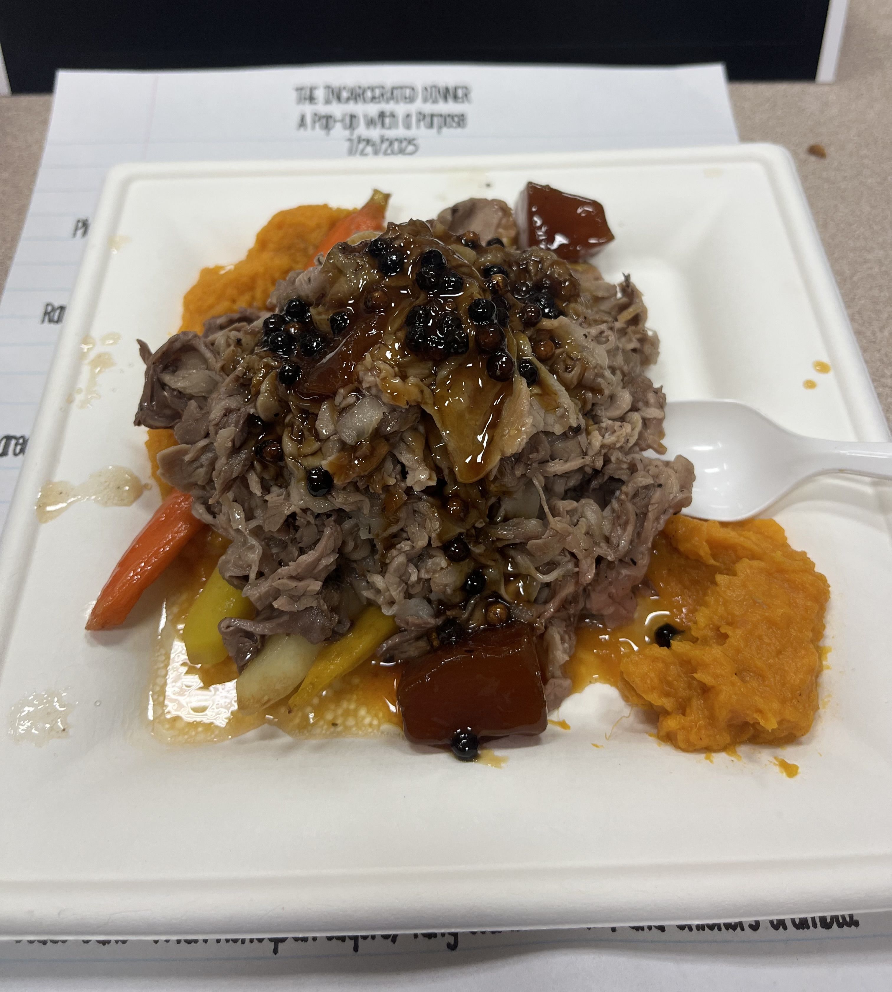 Plate of shredded meat with dark sauce, served over cooked carrots and yellow vegetables with orange mashed side, on a white disposable plate with a white plastic spork.