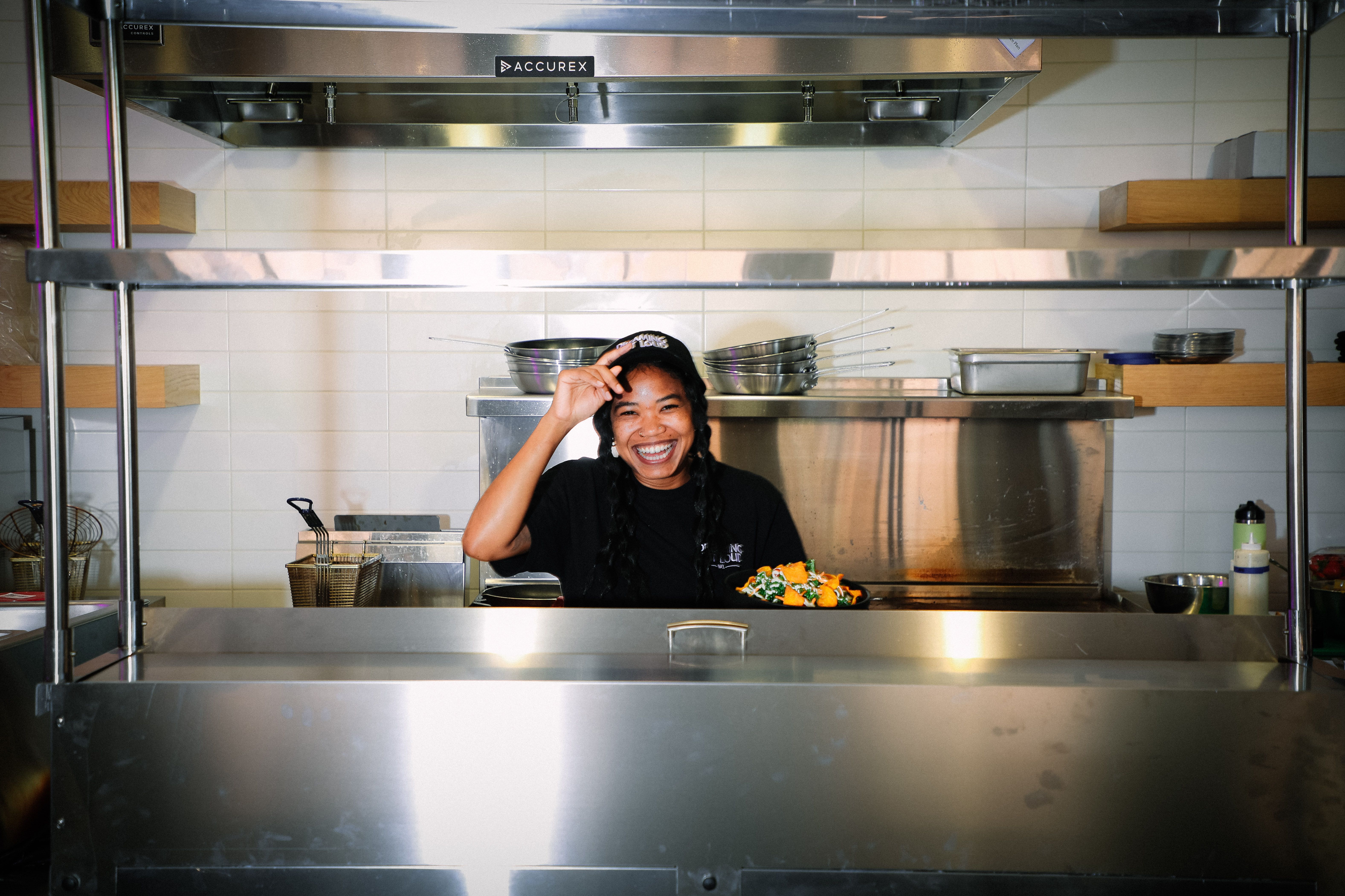 A woman smiles from behind the counter in a kitchen with stainless steel appliances 
