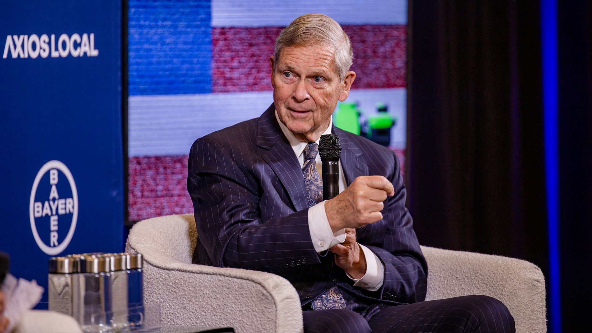 A gray-haired man in a dark pinstripe suit sits in a beige armchair, holding a microphone, speaking on a stage. Behind him are blue Axios Local banners and a colorful screen.