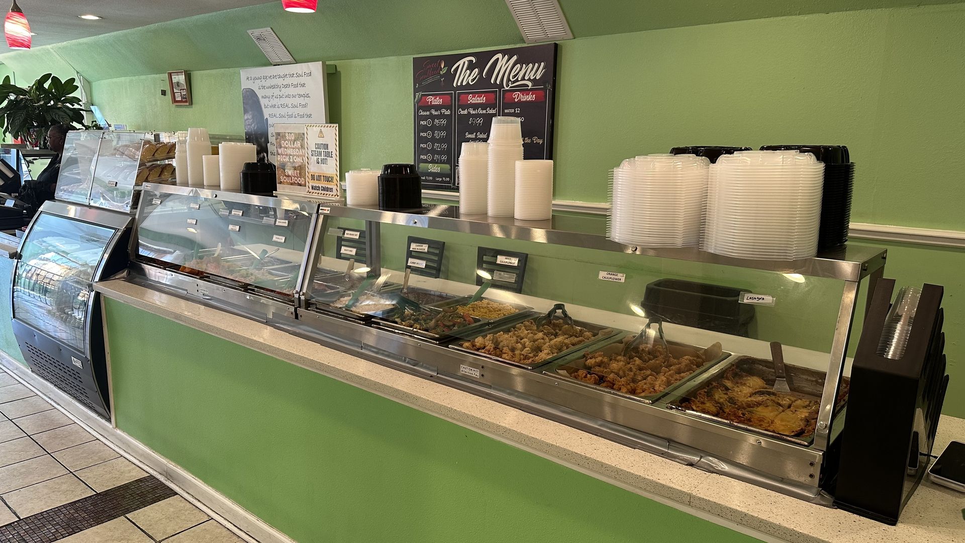 Buffet counter with green walls and various cooked dishes behind glass. Stacks of white and black disposable bowls and plates are on top. A menu board and caution signs are on the wall.