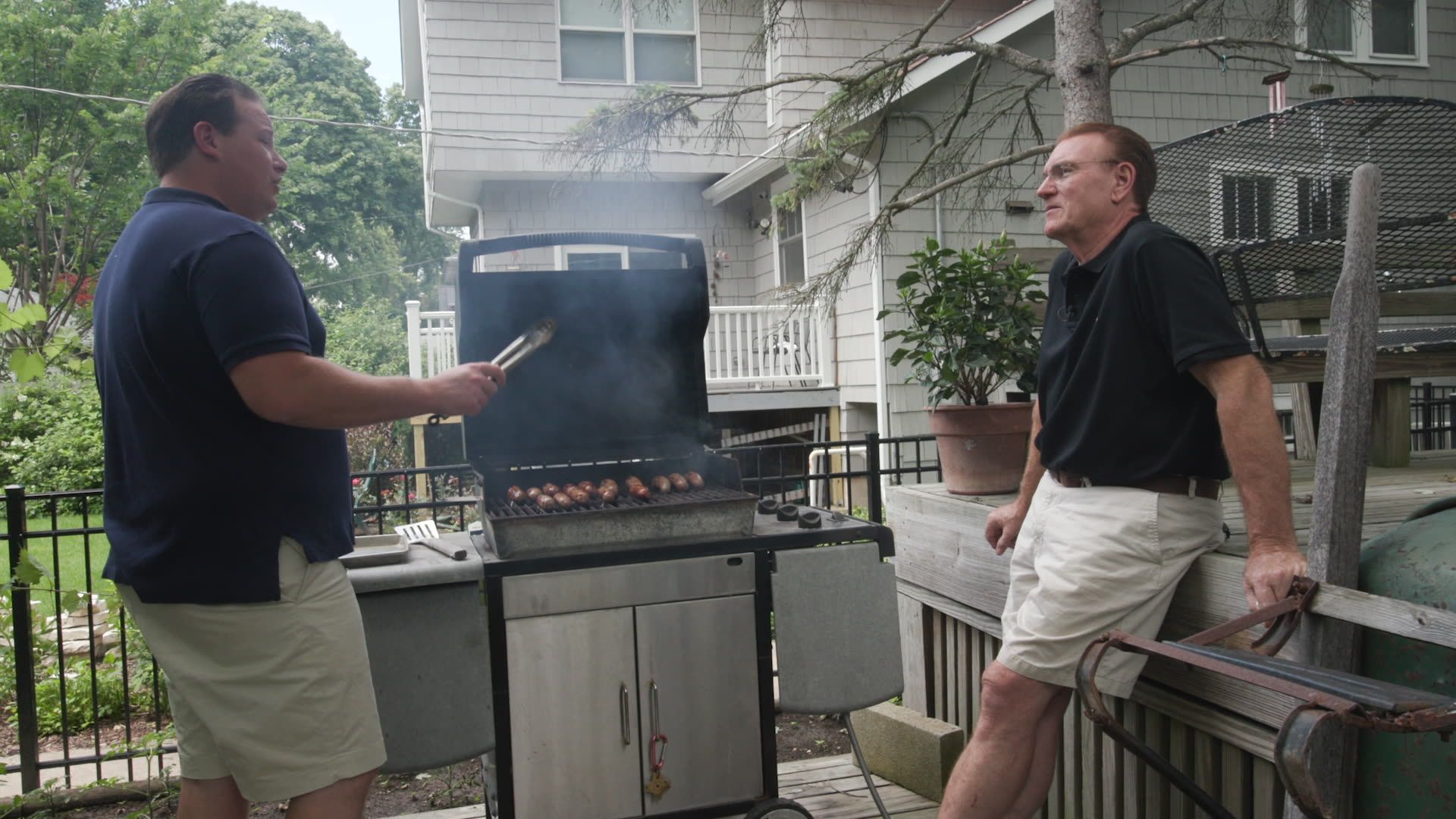 Two men grill sausages in a backyard. Left man in a navy shirt uses tongs at a smoky grill while the other watches; a deck, house, and greenery surround them.