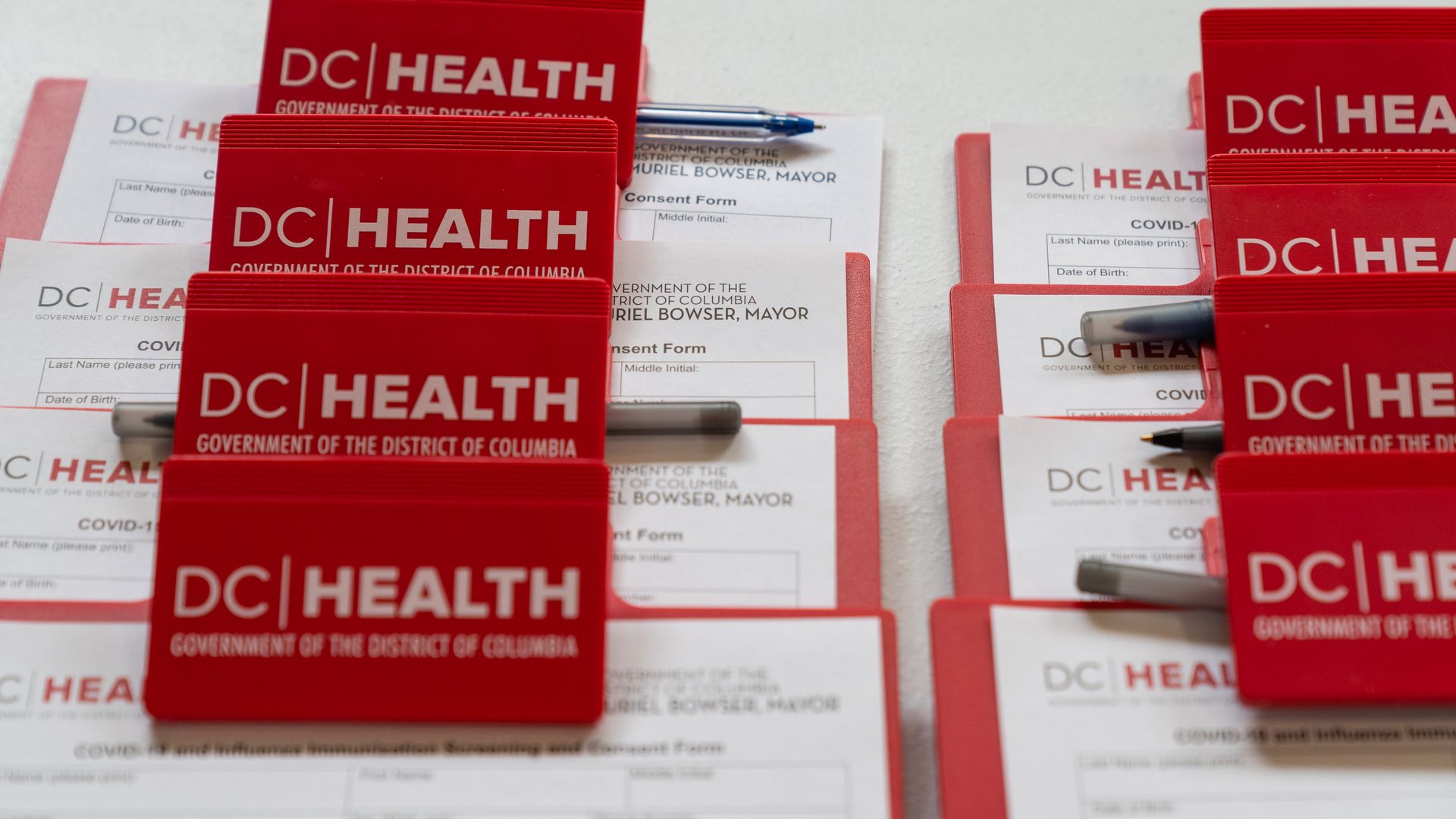 Rows of red folders labeled DC Health with Government of the District of Columbia text, containing COVID-19 vaccine consent forms and pens on a white surface.