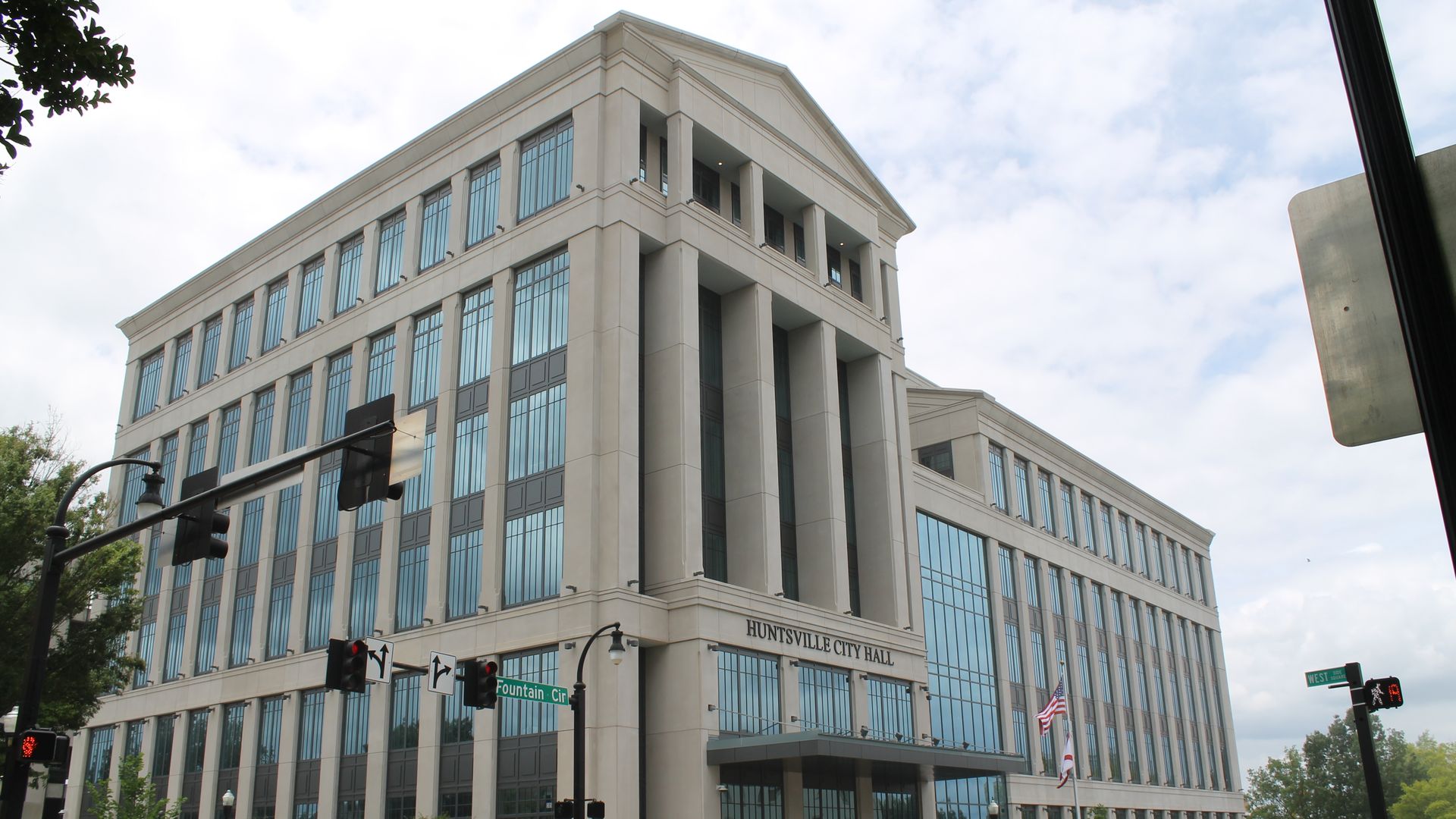 Large modern Huntsville City Hall building with many tall glass windows and beige stone exterior, located at Fountain Circle and West street intersection with traffic lights and flags.