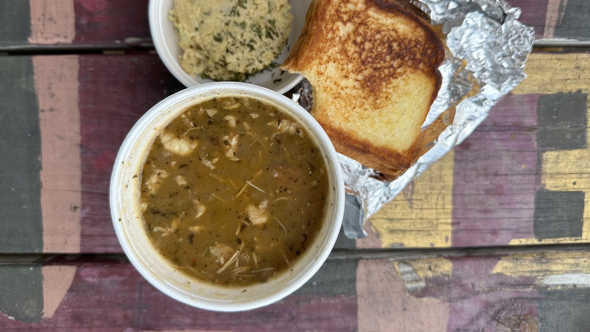 A table is set with a styrofoam bowls of gumbo and potato salad, and a grilled cheese sandwich on a piece of aluminum foil. The sandwich has had a bite taken out of it.