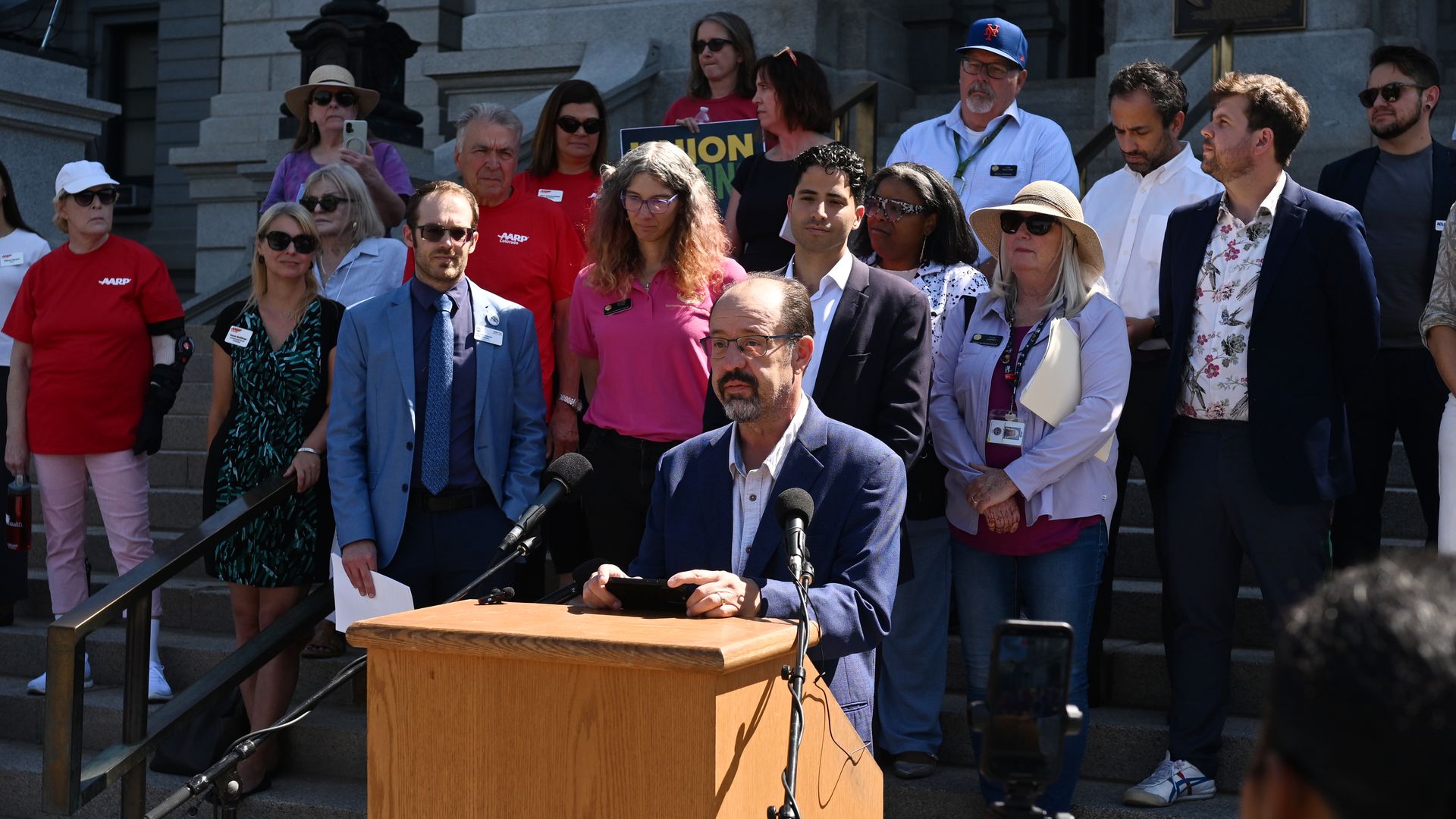 Man in blue jacket speaks at wooden podium with microphones; group of diverse people stand behind him on steps outside stone building, some wearing red AARP shirts and sunglasses.