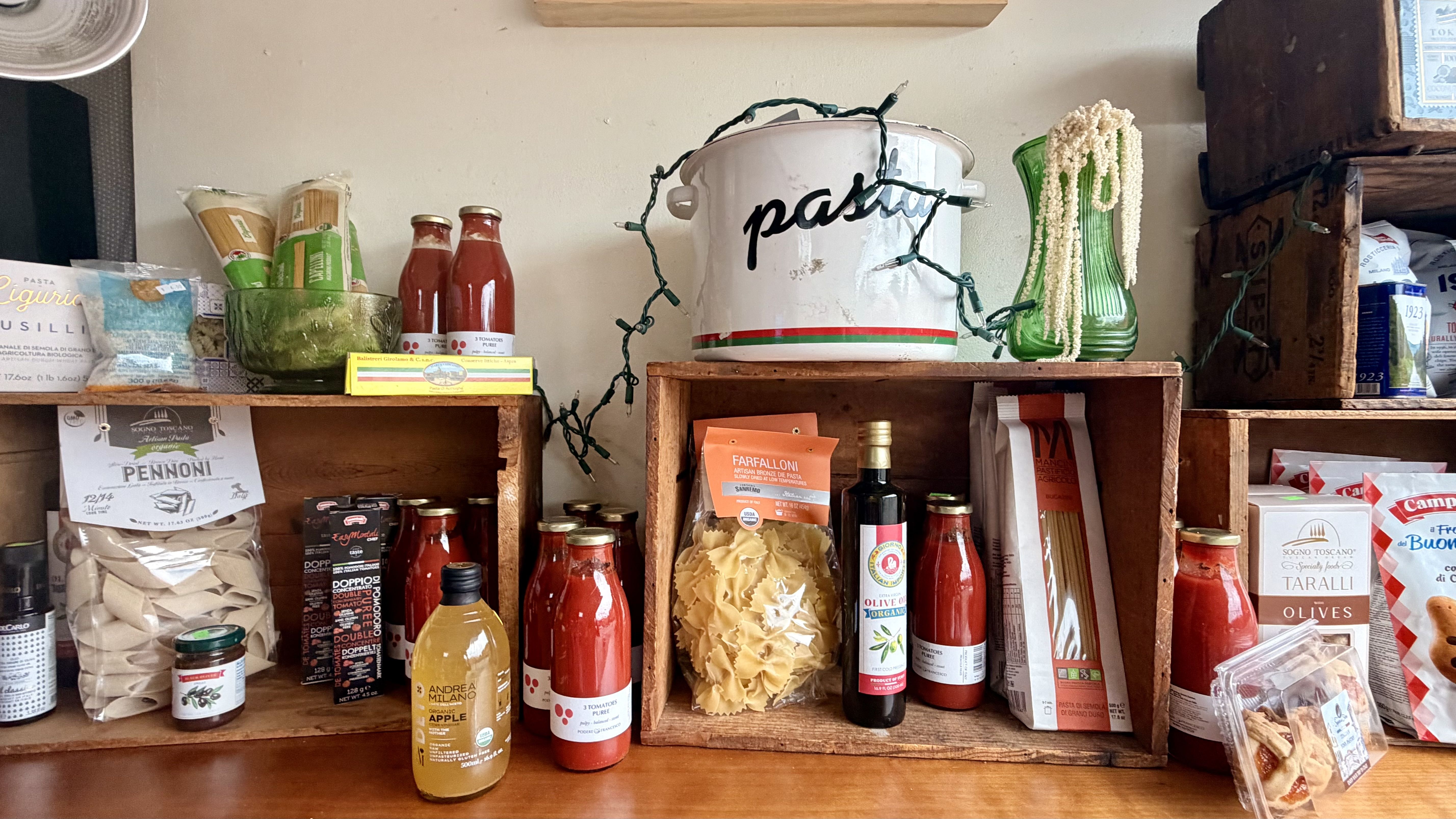 A rustic pantry shelf filled with pasta and sauces: a white enamel pot with the word pasta wrapped in string lights, flanked by a green glass bottle and bags of pasta and jars.