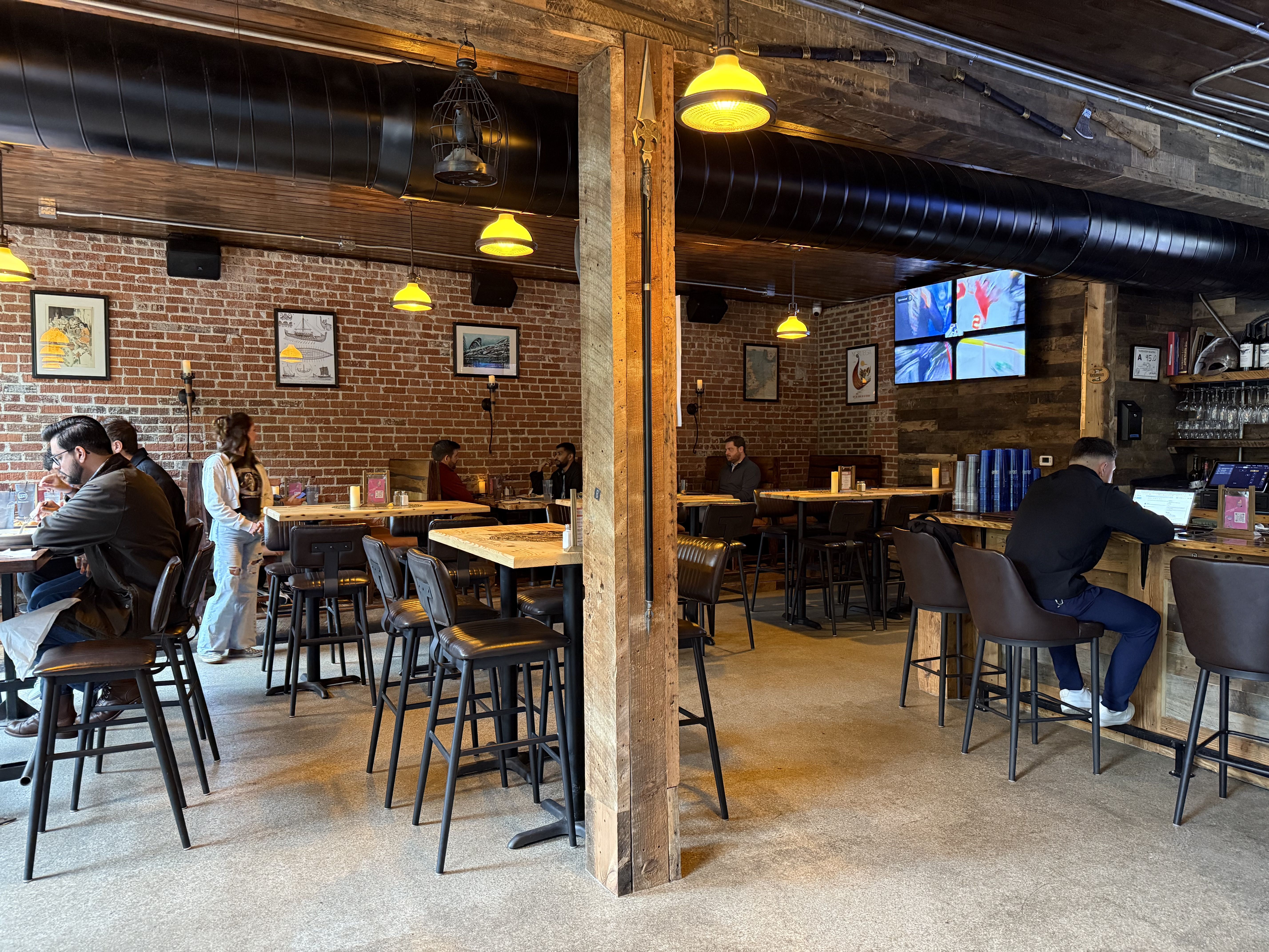 Interior of a rustic cafe with exposed brick walls, wooden beams, and yellow hanging lights. People sit at wooden tables and bar seating, some working on laptops, others chatting.