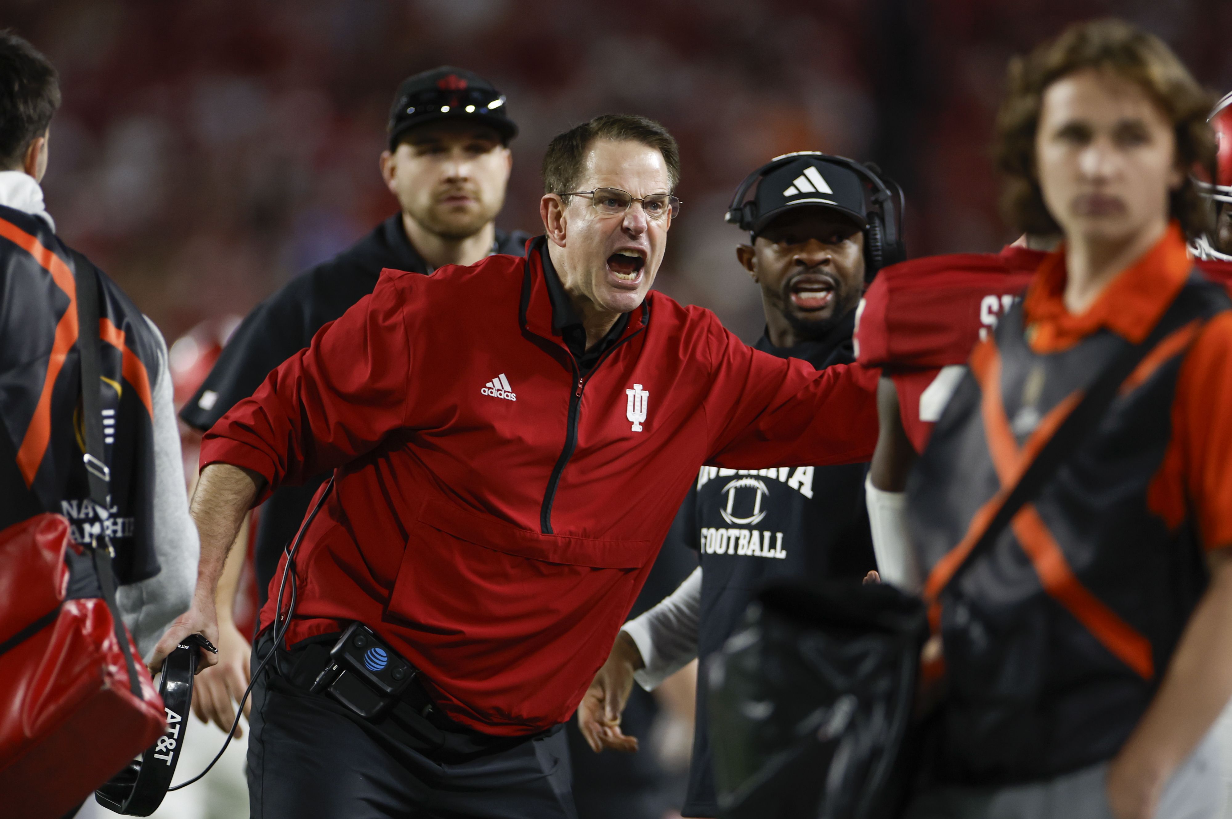Man in red Indiana University jacket shouting, surrounded by coaches and staff on football sideline during game; blurred crowd background.