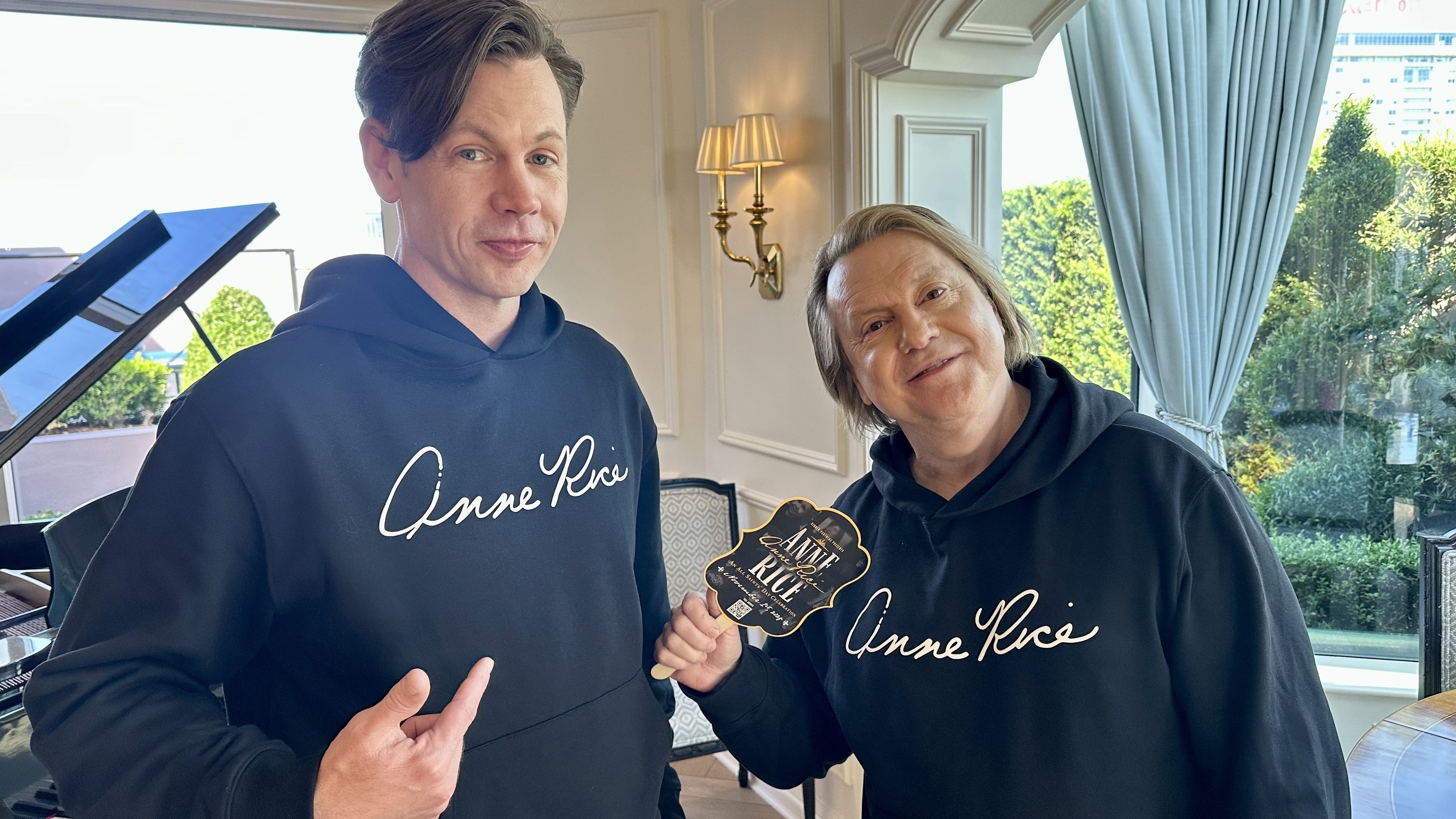 Two people indoors posing for a photo. Left: man in a black hoodie; right: smiling woman in a matching hoodie holding a black-and-gold badge. A room with a piano; greenery outside the window.