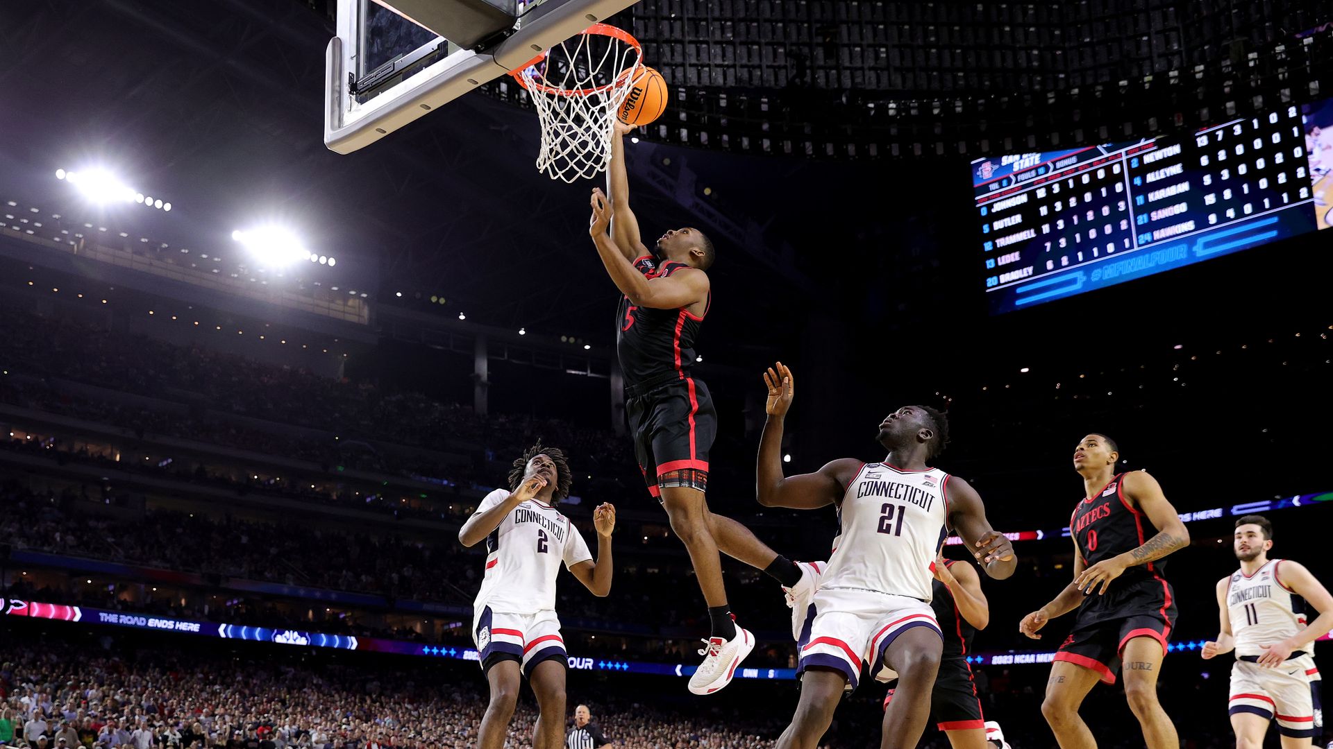 A college basketball player in a black and red jersey drives to the basket for a layup.