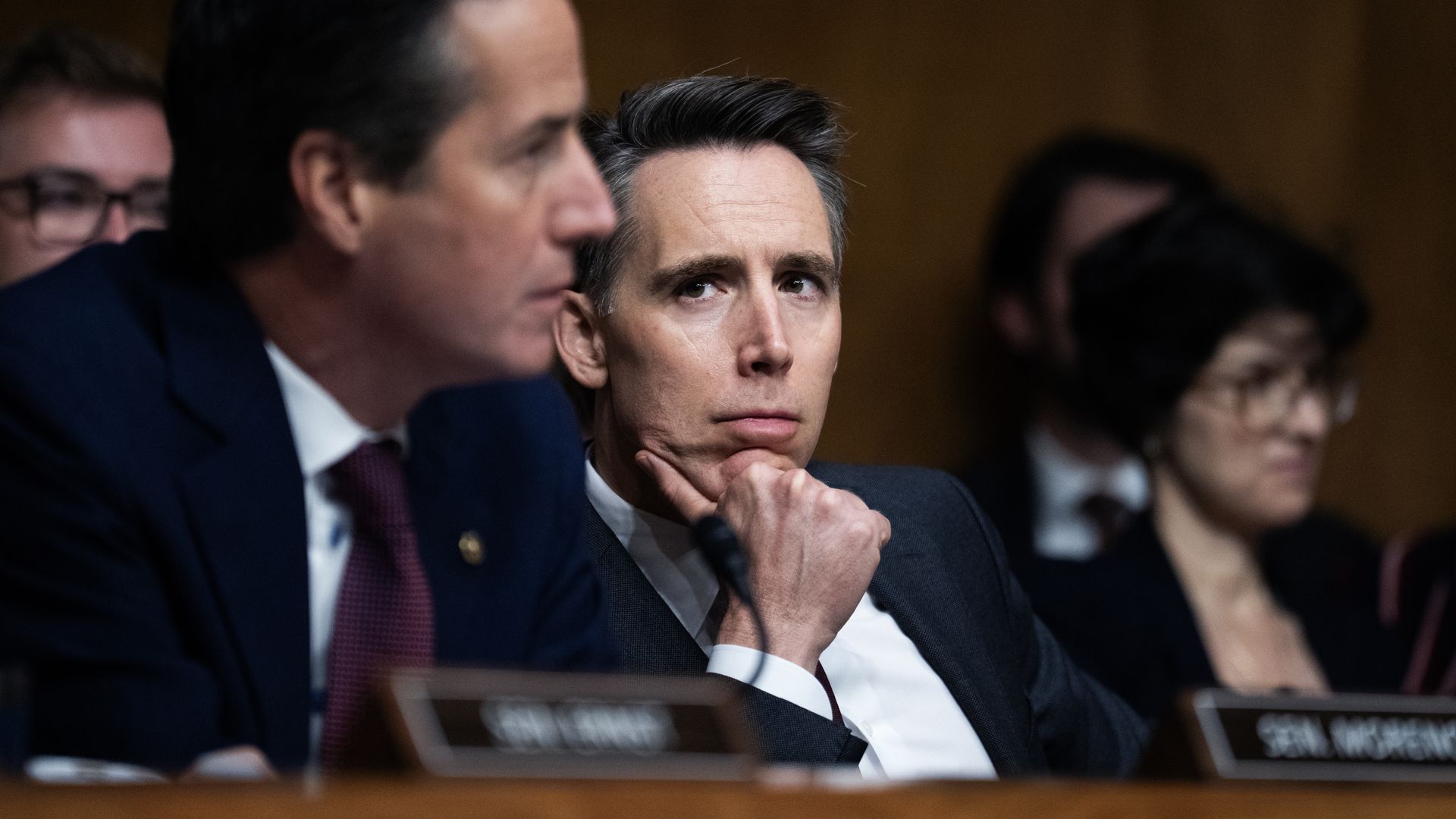 Centered man in a dark suit with white shirt, hand on chin, listening seriously during a formal hearing; blurred colleagues surround him.