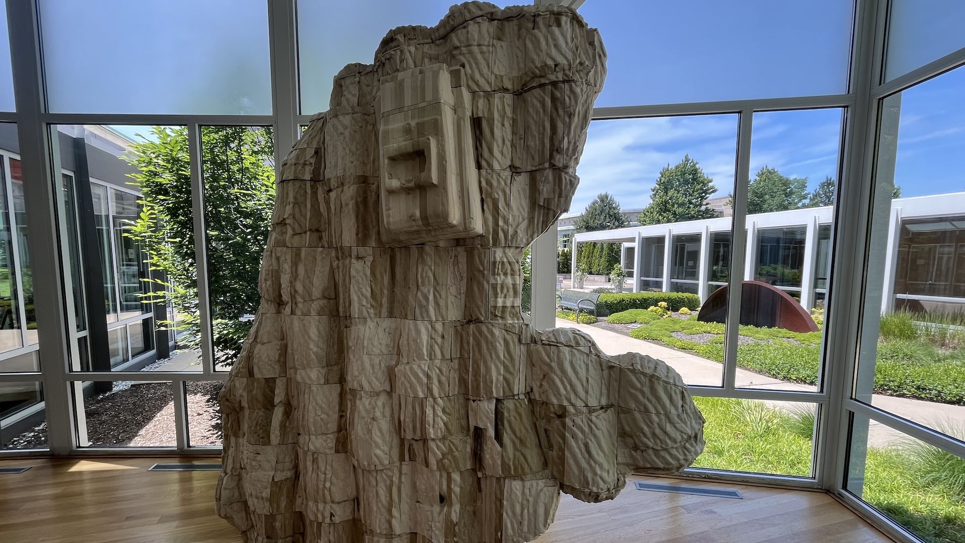 Large beige stone sculpture resembling a layered rock formation stands inside a glass-walled atrium, with an outdoor courtyard, trees, and blue sky visible through the windows.
