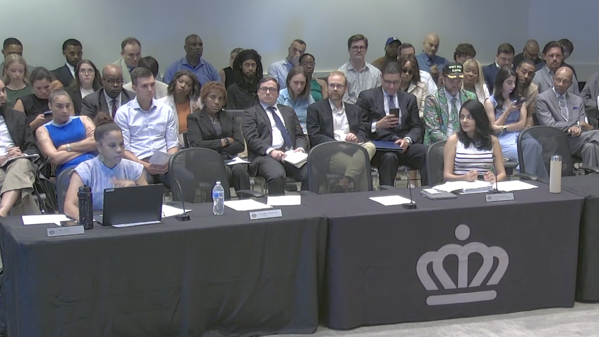 Conference room with a long black-draped table and microphones at the front; a diverse panel sits behind it, facing a seated audience. A crown logo is on the tablecloth.