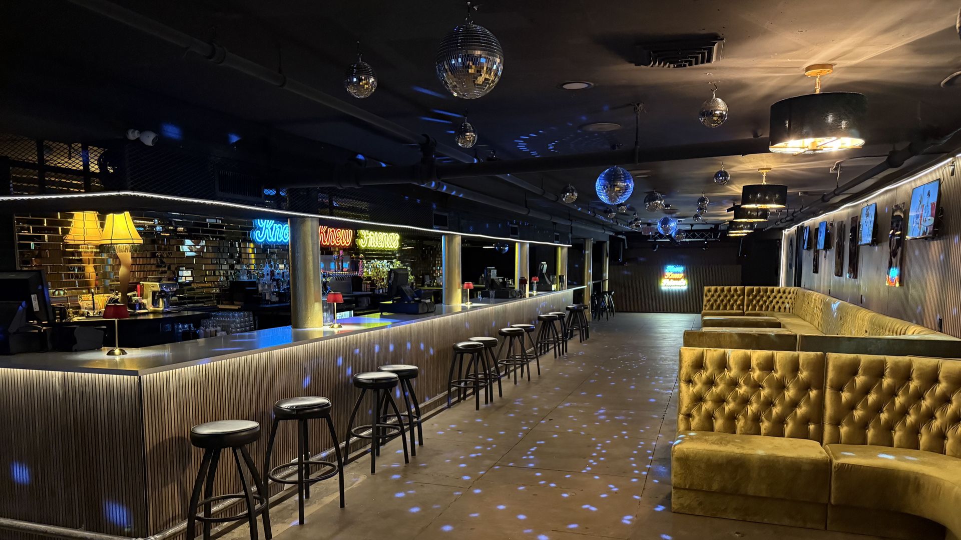 Dimly lit stylish bar with metallic mirrored wall, long bar counter with stools, yellow velvet tufted couches, disco balls on ceiling, and neon signs reflecting blue and red lights.
