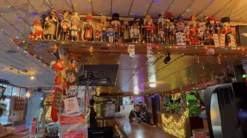 Pan across a festively decorated bar filled with colorful string lights, rows of nutcracker figures on high shelves, and patrons chatting at the counter amid bright holiday ornaments and cozy decor. 