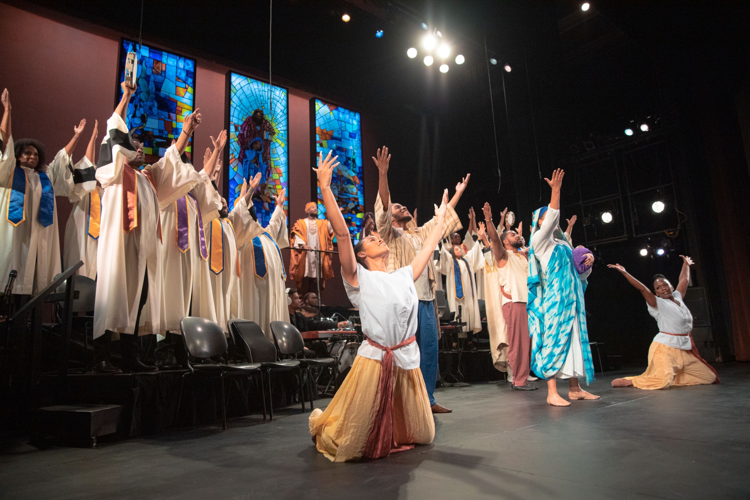 Performers as part of a choir on stage reach their hands up toward the ceiling. 