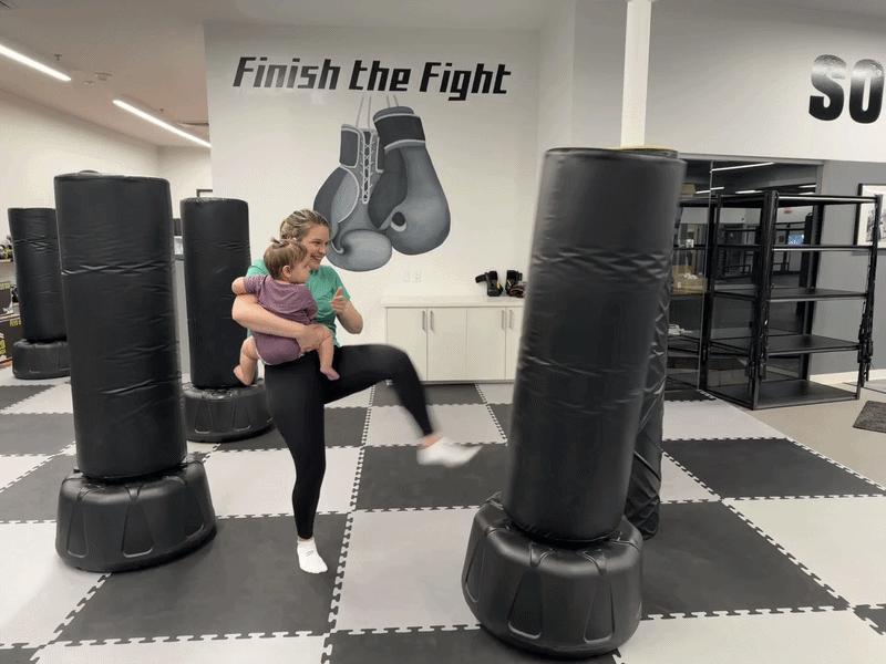 A gif of a woman holding a baby and kicking a heavy bag in a gym.